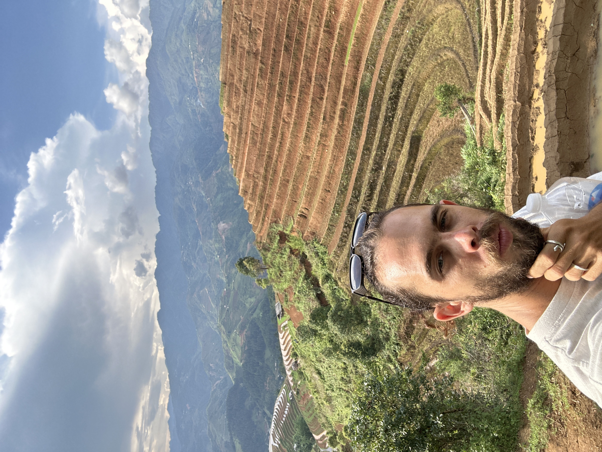 The image, correctly oriented, is a selfie or portrait of a man set against a panoramic view of terraced rice fields in Lao Chai, Vietnam. The man, positioned on the right side of the frame, has a beard and looks directly at the camera with a neutral expression. He wears a white t-shirt and sunglasses are perched on top of his head. Two silver-toned rings are visible on his left hand; one appears to be a crescent or open-ended design. A clear plastic water bottle is partially visible near his hand, though no text on its label is legible.
The background showcases the distinctive, intricate patterns of vast rice terraces carved into the steep hillsides. Some terraces display bare, reddish-brown soil, suggesting they are either freshly prepared for planting or recently harvested. Other sections are covered in lush green vegetation, indicating active growth. Beyond the immediate terraces, a sweeping landscape of verdant mountains stretches into the distance, appearing hazy under a bright blue sky. Large, fluffy white clouds are scattered across the sky, suggesting clear and sunny weather, likely during the morning or afternoon. The scene captures the harmonious blend of human cultivation and natural beauty in a mountainous agricultural region.