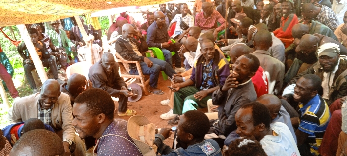 A large group of seated African men are gathered outdoors under a tent-like structure. Many are smiling and engaged in conversation. Some men are wearing jackets and button-down shirts, while others are in casual attire. The setting appears to be a community event or meeting, with chairs and stools used for seating. The overall atmosphere is relaxed and convivial.