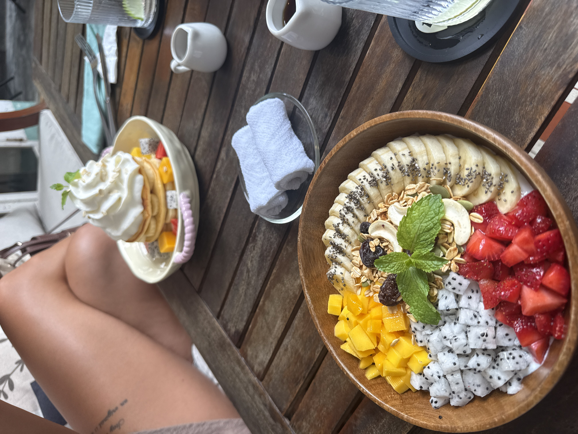 A high-angle shot showcases a wooden table setting. On the table are two food bowls: one with layered crepes, fruit, and whipped cream, and another with yogurt, banana slices with chia seeds, granola, mango, pitaya, strawberries, and a mint garnish. Also on the table are two ceramic mugs, a glass with a drink and cutlery, a small tray with rolled towels, and a cup of liquid. The lower portion of a person is visible, with a tattoo on their leg. The environment suggests an outdoor dining setting. The food style and the setting hint at a health-conscious or brunch-style establishment. The location could be in the city of Đà Nẵng, Vietnam, based on the potential food items.