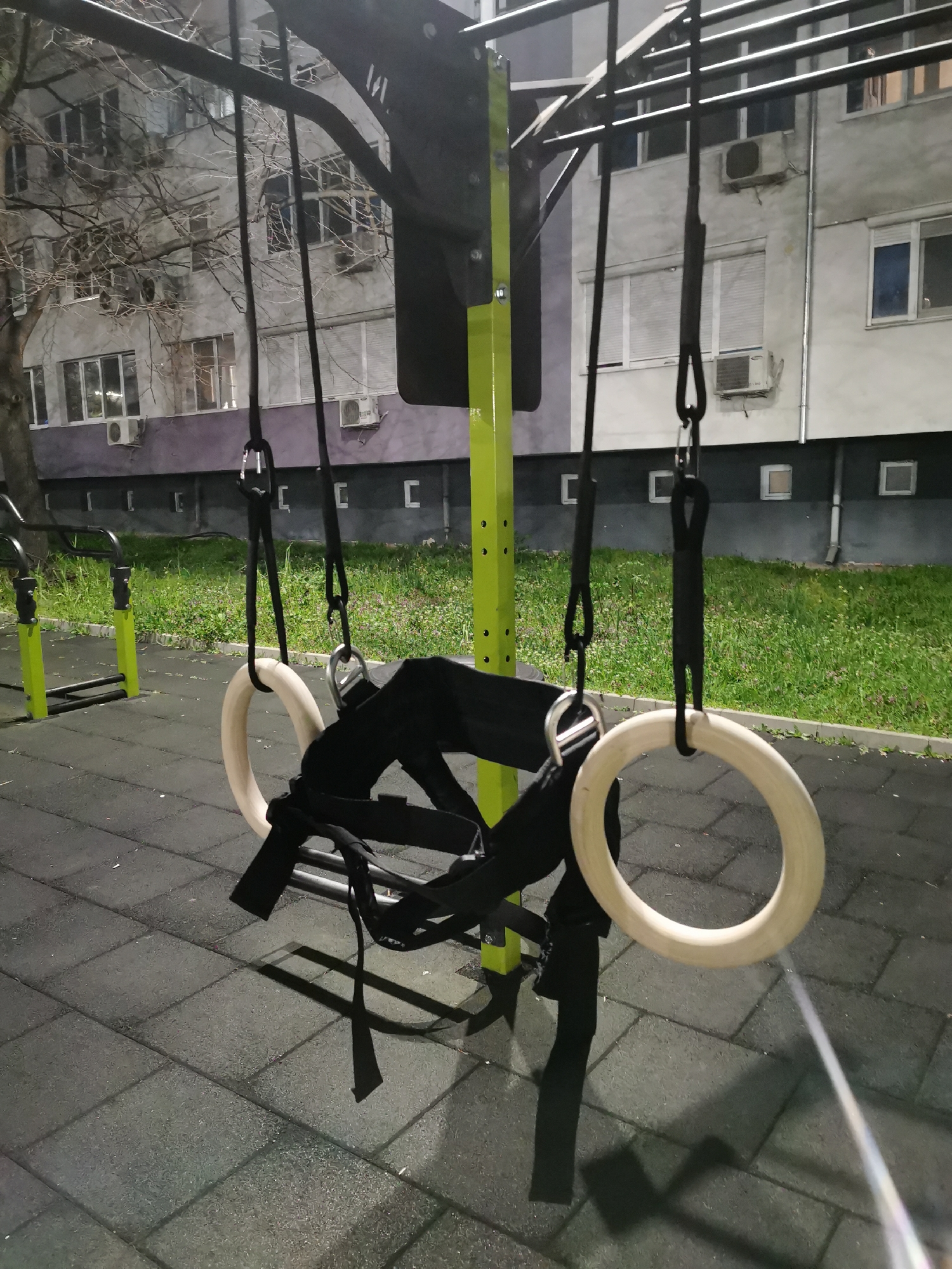The image shows a set of gymnastic rings hanging from an outdoor exercise structure. The rings are wooden, and the straps suspending them are black. The structure is made of dark metal bars and a lime green pole. A black harness or support system is attached to one of the rings. The ground is covered with dark grey rubber tiling. In the background is a multi-story building with multiple windows, some with air conditioning units. There is a tree without leaves and some grass present. The photograph was taken at night. The scene is likely a public park or outdoor workout area, possibly in Burgas, Bulgaria.
