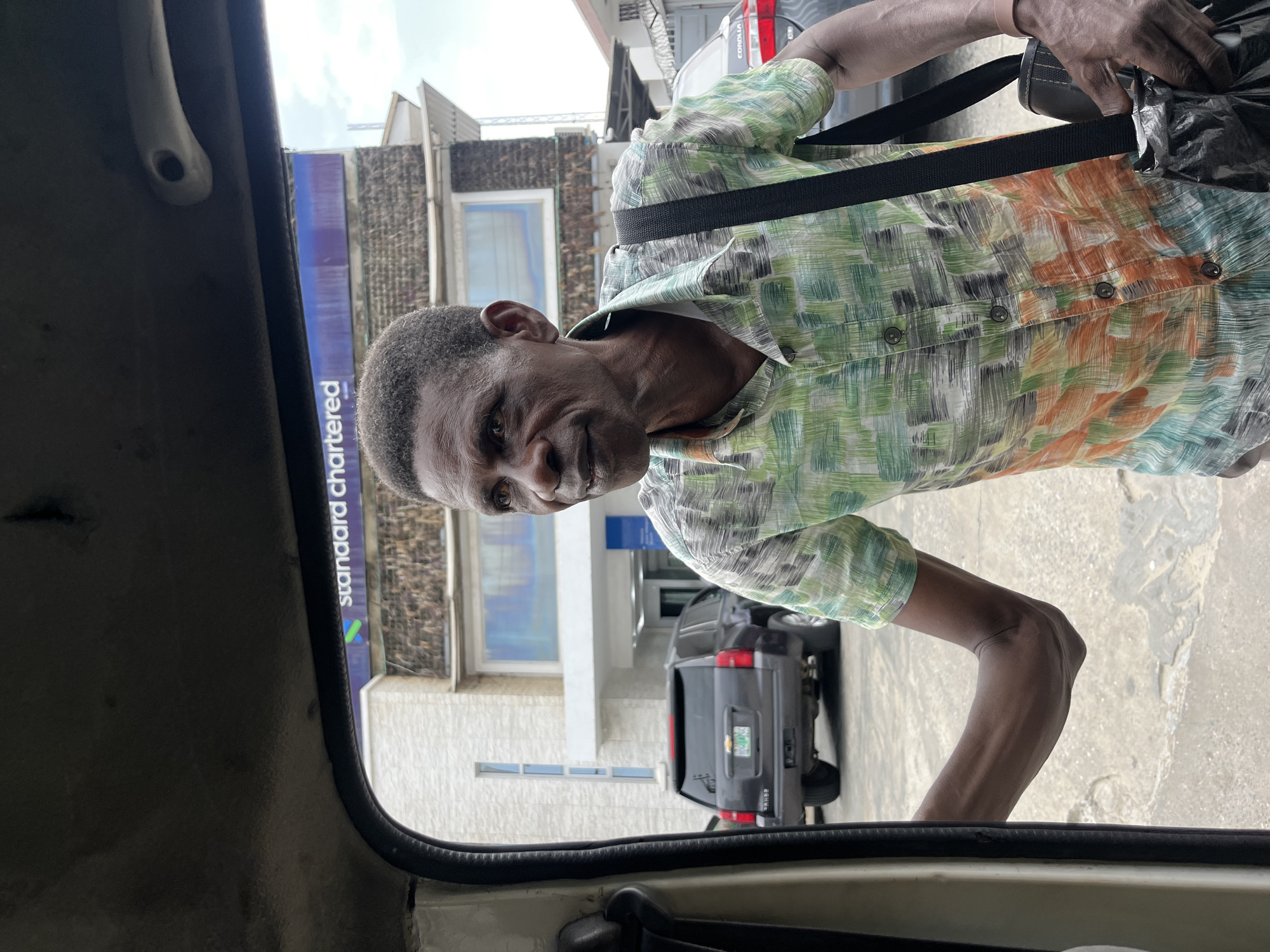 A medium shot taken from inside a vehicle in Itirin, Nigeria. In the foreground, the interior of the vehicle is visible, including the door frame. Through the window, an older, dark-skinned man stands on the sidewalk, wearing a green and orange button-down shirt and a black shoulder bag. In the background, a Standard Chartered bank building is visible, along with a dark SUV parked nearby. The environment suggests a street scene in a commercial area, possibly a business district within Itirin. The scene is captured during daylight hours, with a partially cloudy sky.