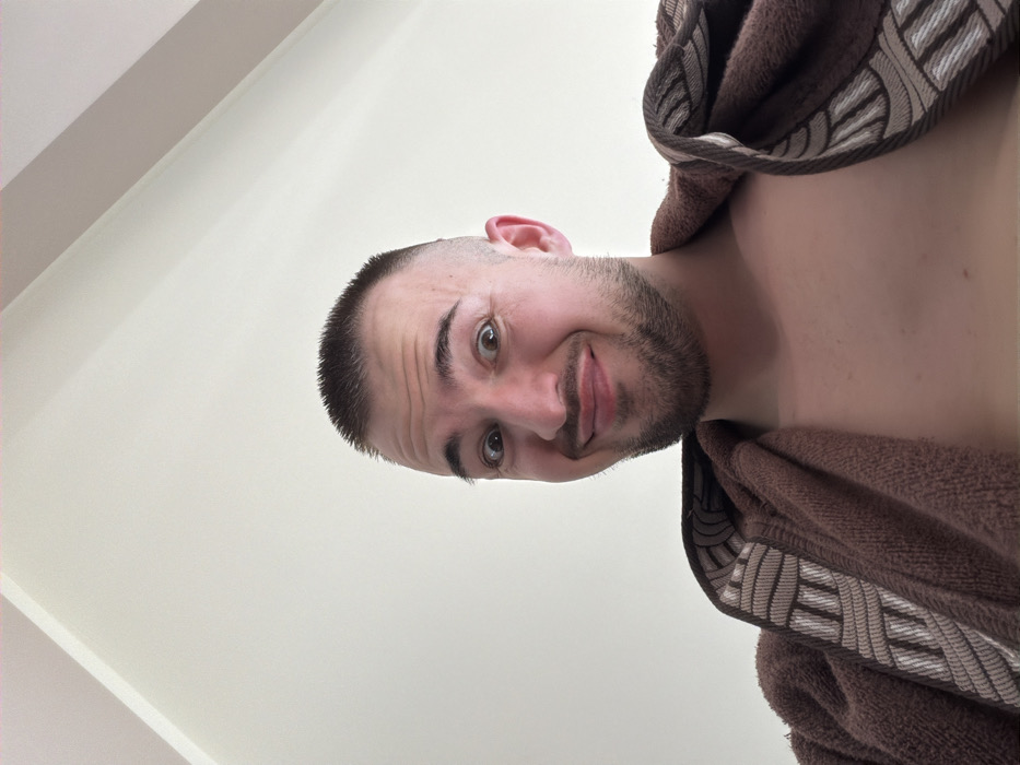 A low-angle, forensic portrait depicts an adult male, estimated 20-30 years of age, situated indoors in Kladnitsa, Bulgaria. The subject presents with light skin, a short buzz cut hairstyle, and light facial stubble. His eyes are wide-open, pupils centered, and his mouth is set in a slight, pursed smile directed towards the camera. He wears a brown terrycloth robe draped open, revealing his bare chest; the robe's collar features a distinct geometric pattern in beige, white, and brown. The background is an undecorated, light-colored (off-white) interior wall and ceiling, with a visible corner junction in the upper left. The subject's head is slightly cropped at the crown.