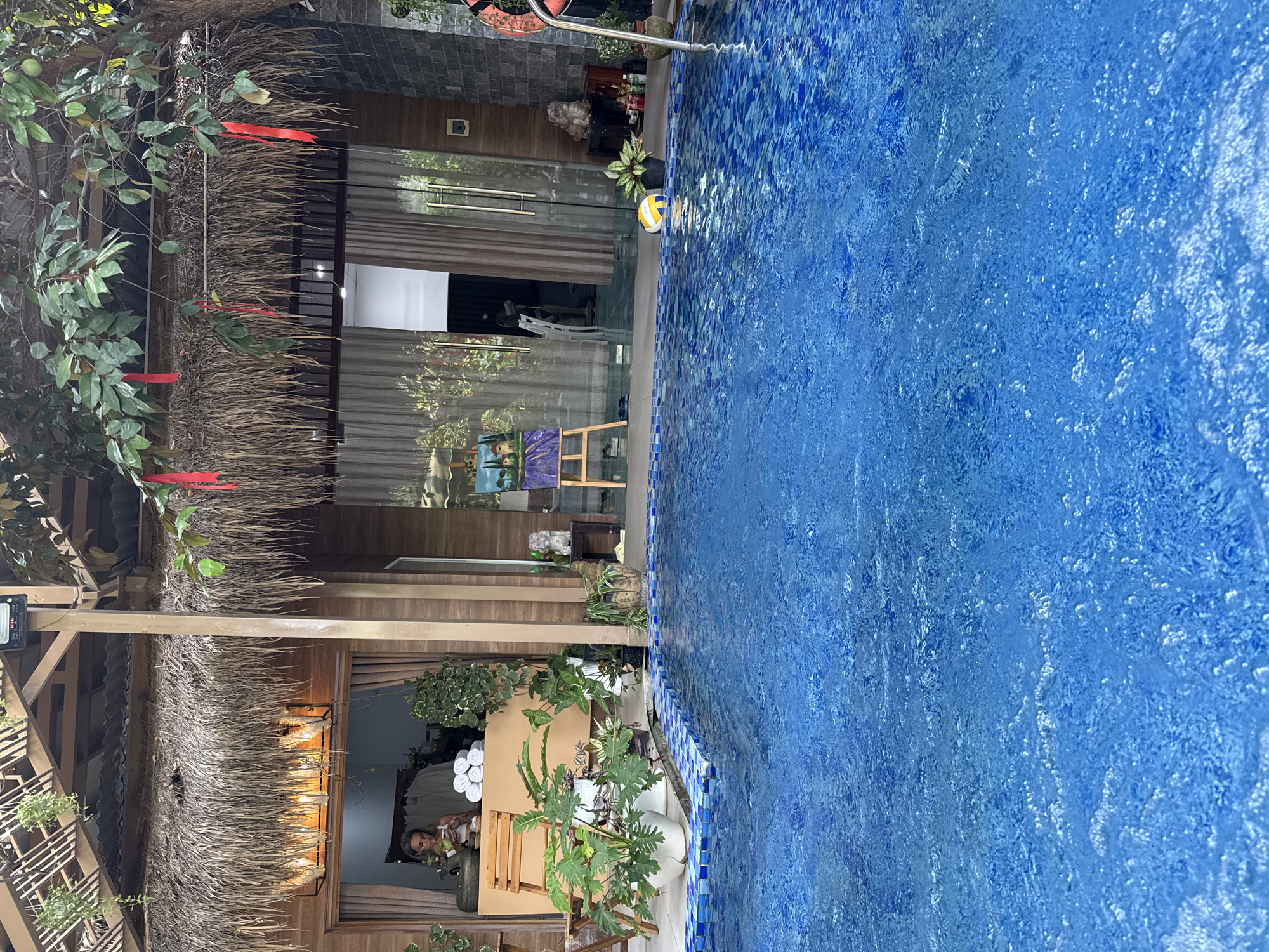 The scene shows a building with a thatched roof next to a blue-tiled swimming pool, likely located within the Bac Natural Heritage Area Trang, Vietnam. The building has a large glass door. An easel with a painting stands inside the building near the pool edge. A person is seated at a table inside, next to a stack of white towels. Plants are arranged around the table. A volleyball floats in the swimming pool. Red ribbons are tied to the thatched roof structure.