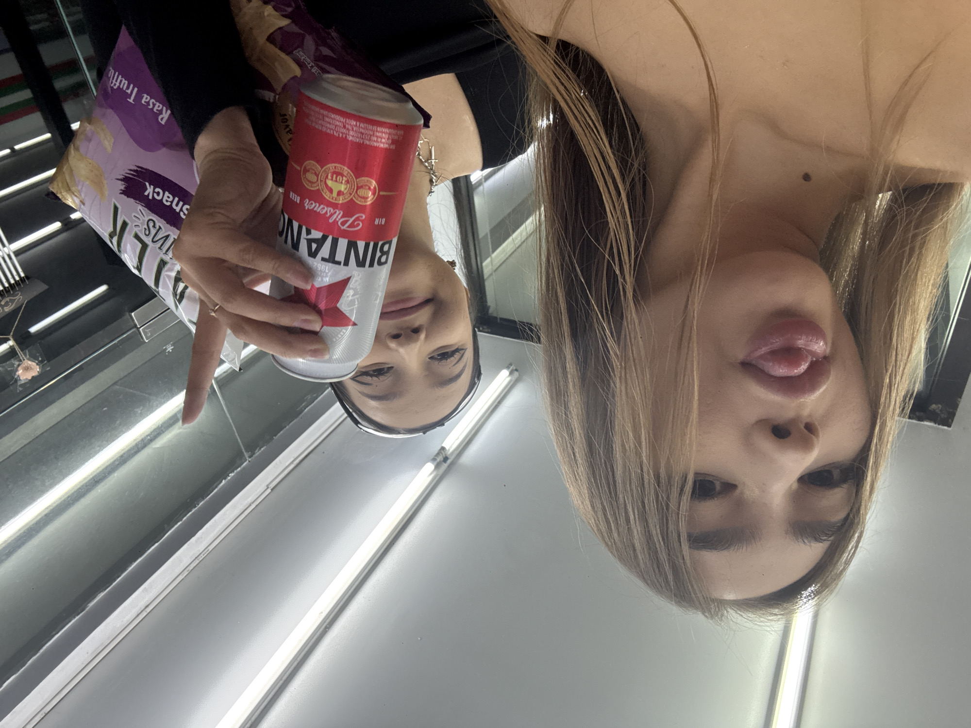 A photograph, taken at close range, shows a woman holding a can of Bintang pilsener beer and a bag of potato chips (Kriuk Rasa Truffle) in a store. She is looking at her reflection. The beer can label indicates "BIR Pilsener" and the date "1952", which refers to when the brand was established. The bag of chips notes "Rasa Truffle," a flavor, and gives its weight as 60g. The store's lighting features fluorescent tubes running parallel to the shelves. This photo was taken in North Kuta, Indonesia.