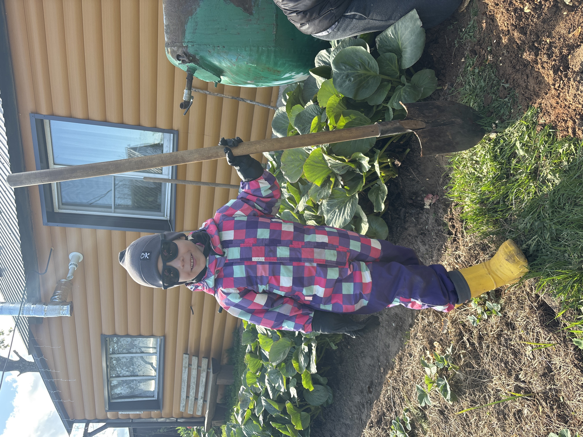 A child stands in a garden with a shovel in a location identified as Ваньки, Russia. The child wears sunglasses, a gray beanie, a colorful checkered jacket, purple pants, and yellow boots, along with black gloves. The child holds the shovel upright. Behind the child are large green plants, a green water tank, and a brown wooden building with windows. The ground is a mix of soil and grass.