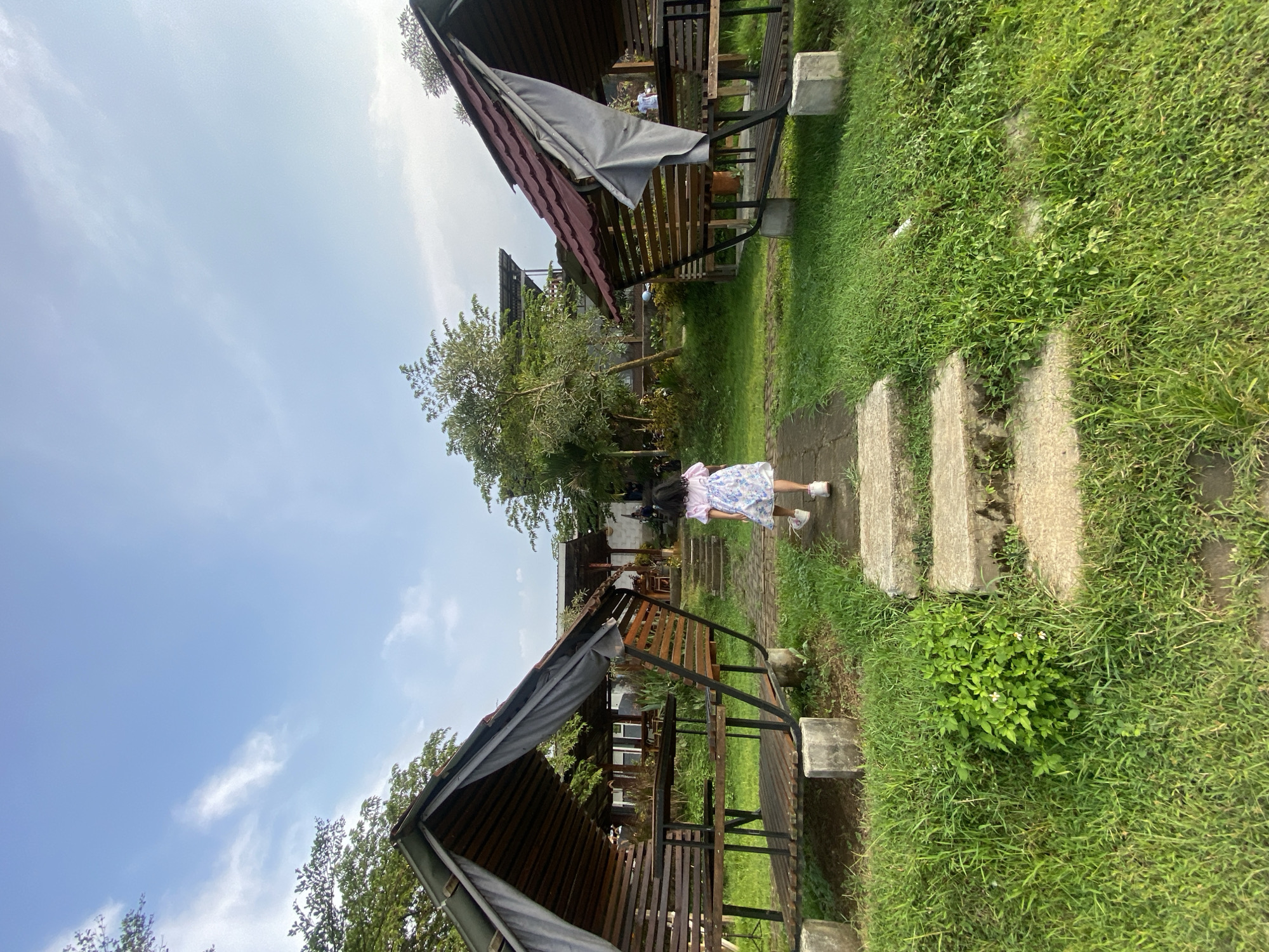 A young girl walks on a stone path between two wooden structures with roofs in Penanggungan, Indonesia. The structures are surrounded by grass and trees under a partly cloudy sky.