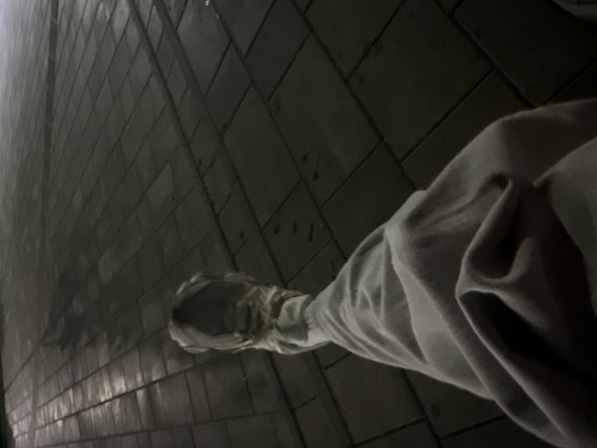 The image shows a person's leg and foot wearing a light-colored sweatpant and a white sneaker. The person is sitting on a tiled surface, which appears to be a walkway or plaza. The lighting suggests it is nighttime or a dimly lit indoor space. The tiles are dark and laid in a rectangular pattern. A shadow is cast on the tiles from the person, stretching diagonally across the frame. The environment is not clearly identifiable as being in Dnipro, Ukraine, as there are no specific landmarks or identifying features of that city visible.
