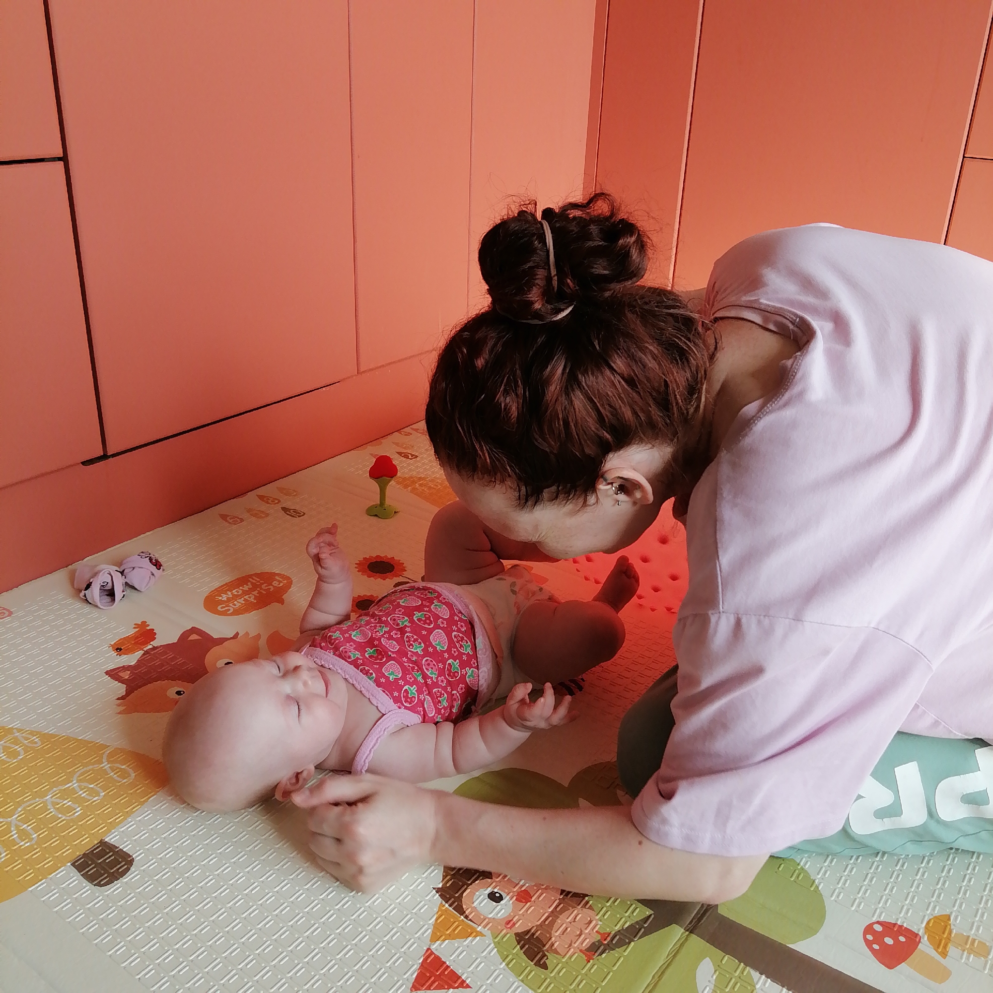 In Trubacheyevka, Russia, a woman with dark, reddish-brown hair tied in a messy bun, wearing a light pink shirt, is positioned on a playmat. She is leaning forward, appearing to kiss the forehead of a baby lying on their back. The baby, wearing a pink onesie with a strawberry pattern and a diaper, has their eyes closed and appears to be smiling. The playmat features colorful cartoon animals and mushroom illustrations. To the left of the baby are a pair of small pink socks with white polka dots. Behind the woman and baby are floor-to-ceiling peach-colored cabinets. The overall environment appears to be a home nursery or play area.