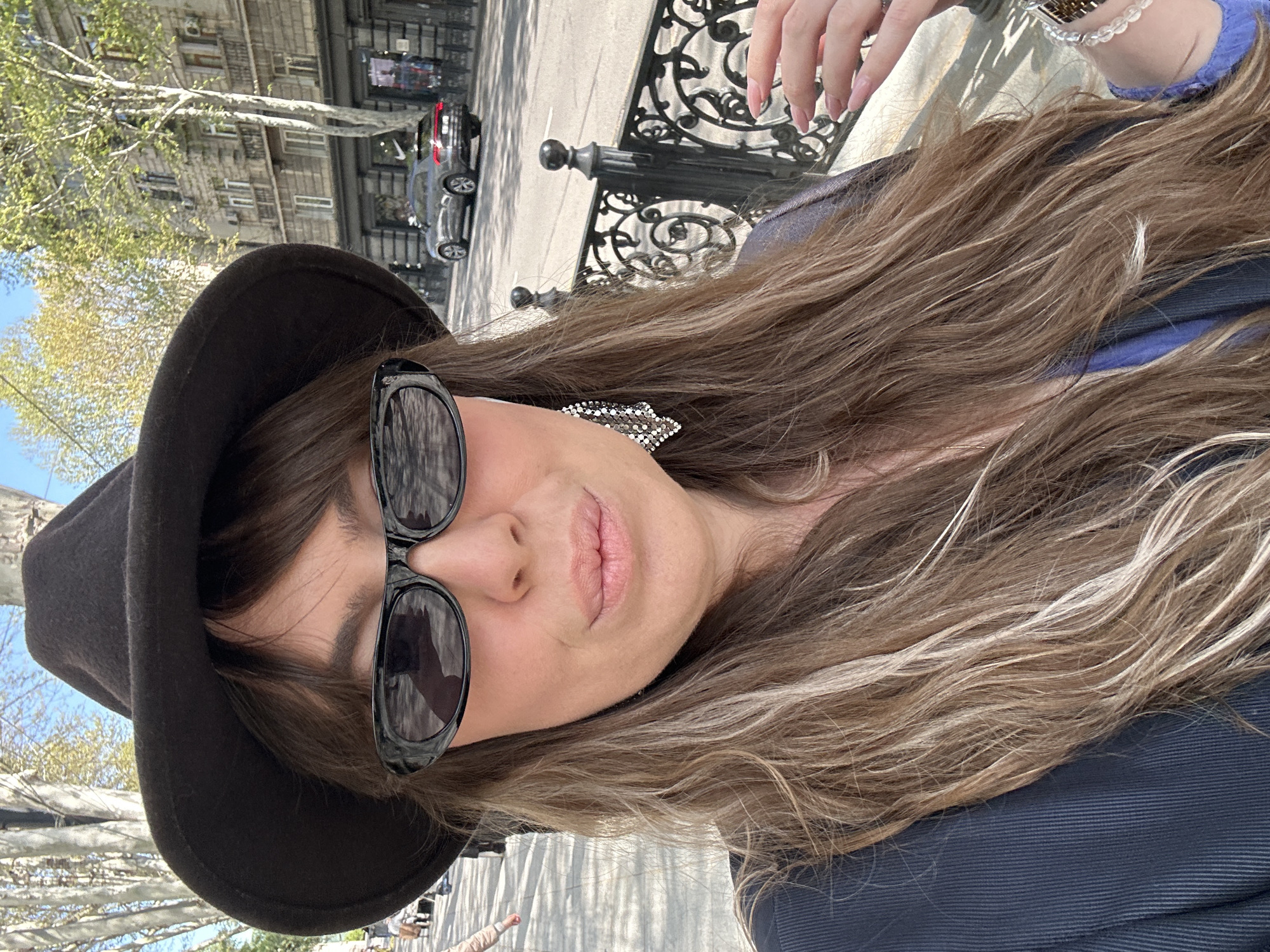 This outdoor portrait captures a woman in an urban setting, likely taking a selfie given the close-up, high-angle perspective. The image is rotated clockwise, making the subject appear on her side.
The main subject is a woman with long, wavy, light brown hair streaked with blonde highlights. She wears large, black sunglasses and a dark-colored (likely black) wide-brimmed hat. Her visible features include her mouth, accentuated with light pink lipstick, and part of her cheek and chin. A sparkling, silver-toned earring with an intricate design is visible on her right ear. She is wearing a dark blue garment, possibly a jacket or top, with visible fabric texture. Her right hand, featuring long, light-colored fingernails and a beaded bracelet, is visible in the upper right corner of the frame.
The background reveals an urban environment on a sunny day. A blue sky is visible at the top. Behind the woman, there's a street with a dark-colored car, possibly a sedan, parked or moving. Buildings with multiple windows and air conditioning units line the street. Sparse trees with light green, budding leaves suggest springtime. A decorative black wrought-iron fence or railing is also present, adding to the city aesthetic. The strong sunlight casts clear shadows, indicating bright daylight. There is no visible text in the image.
The overall scene suggests a casual outing in a city like Tbilisi, Georgia, as indicated by the context.