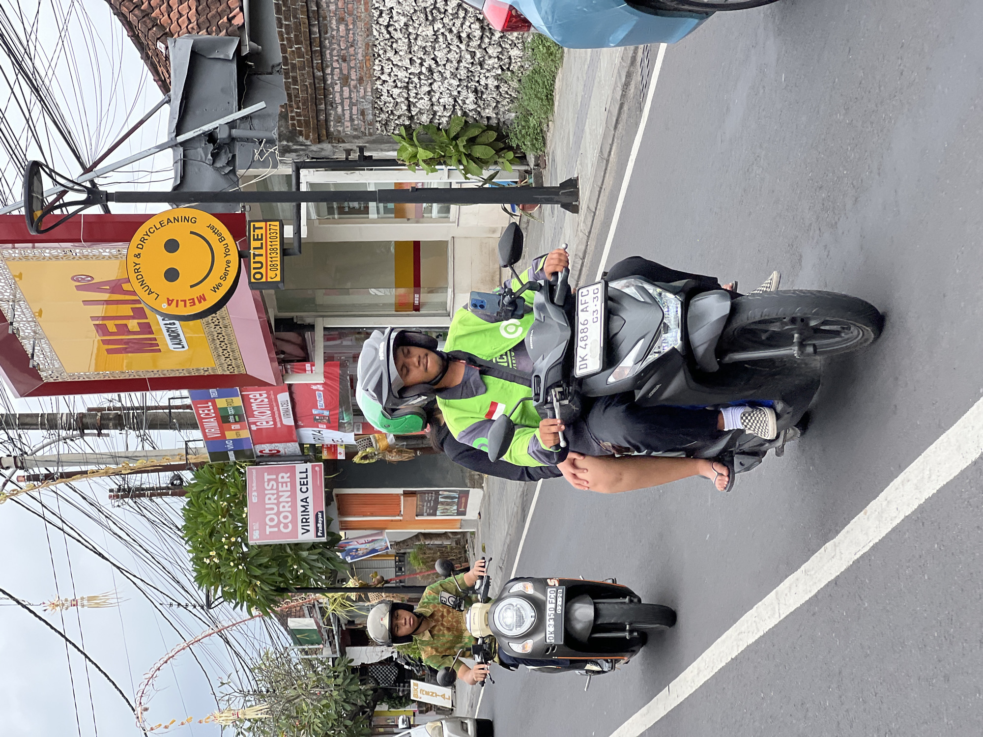 A medium shot shows two people riding a scooter on a street in Denpasar, Indonesia. The scooter is dark gray with a license plate that reads "DK 4886 AFC". The driver is wearing a green jacket and a helmet, and the passenger is wearing a helmet and a green shirt. The scooter is on the right side of the road, and there is a blue car to the right of the scooter. In the background, there are buildings with signs for "Melia Laundry & Drycleaning" and "Virima Cell". The sky is overcast.