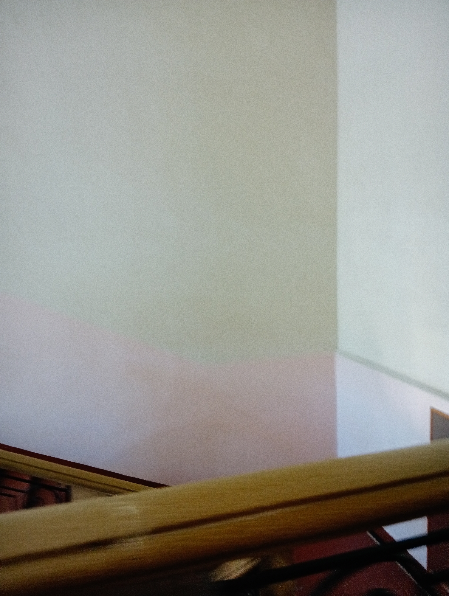 The image displays an interior architectural scene, likely within a building in Poltava, Ukraine. The primary subjects are the walls and a prominent handrail. The walls exhibit variations in light color, with a large section appearing light greenish-grey, meeting a lighter, almost white wall at a sharp vertical corner on the right. A lower part of the wall on the left transitions into a muted light pink or subtle purple hue.
A light brown wooden handrail extends diagonally across the lower foreground, from the bottom left to the mid-right of the frame. Beneath this wooden element, dark metal balustrade details are partially visible, set against a dark reddish-brown background. The scene is brightly lit, creating subtle gradations and perceived color shifts on the wall surfaces. The entire image has a noticeable fine grainy texture. There are no people, activities, or visible text present. The indoor setting does not provide specific cues for the time of day or weather.