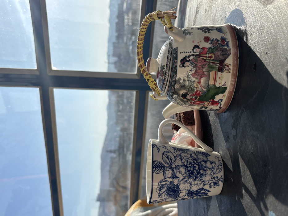 The image displays an indoor scene with a panoramic view of the city of Krasnoyarsk, Russia, from an elevated position. In the foreground, a dark, textured table surface features a white ceramic mug with a blue floral pattern (peonies/roses) on the left. To the right of the mug, a white ceramic teapot with a light brown base is decorated with traditional Asian artwork depicting multiple female figures in colorful robes. The teapot has a yellow woven rattan handle. Situated behind the mug, on a small pink plate, are two dark brown baked goods and one small white, paper-wrapped item. Direct sunlight streams from the right, casting distinct shadows of the mug and teapot onto the table. The background reveals the cityscape through a multi-paned window with dark frames. The urban sprawl of Krasnoyarsk is visible, featuring various buildings, including a prominent dome or steeple structure in the mid-ground. A range of hills or mountains, possibly snow-capped, form the distant horizon beneath a clear, bright blue sky. No individuals are present in the scene.