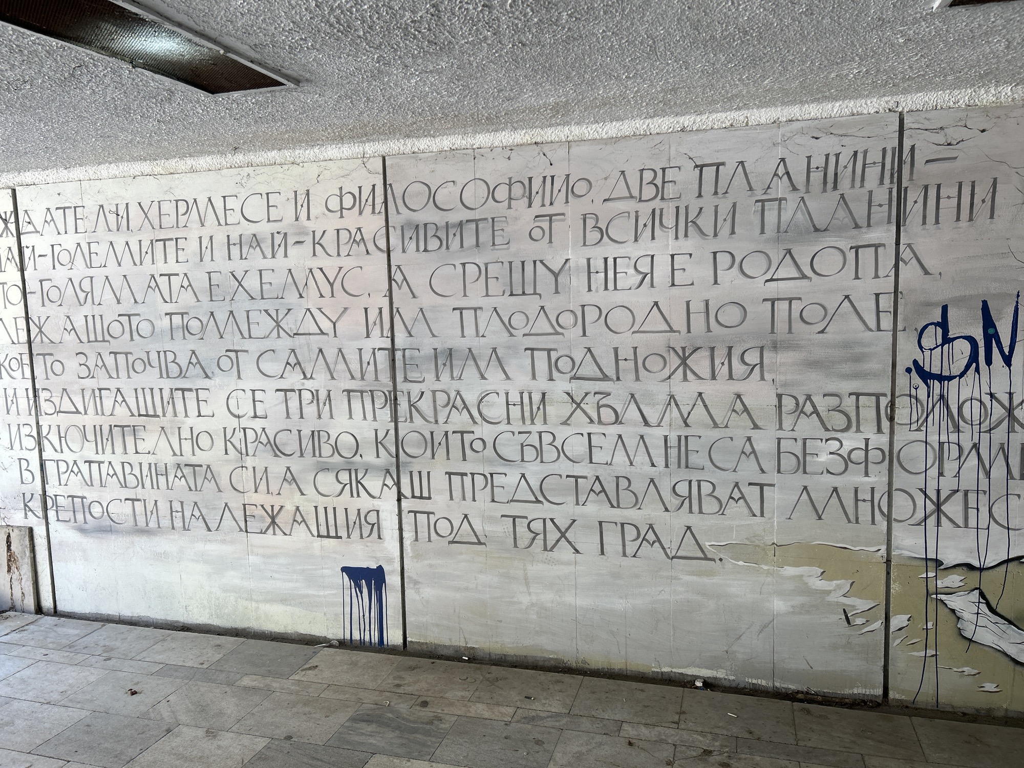 The image depicts a section of a wall, likely indoors, with engraved Bulgarian text. The text appears to be part of a longer narrative or historical description, potentially related to the city of Plovdiv.
Several sections of the wall are defaced with graffiti, including blue paint dripping and an abstract scribble.
The wall is attached to a paved floor. The environment suggests an urban, potentially abandoned, space. The lighting is dim, indicating either indoor conditions or a shaded outdoor area.