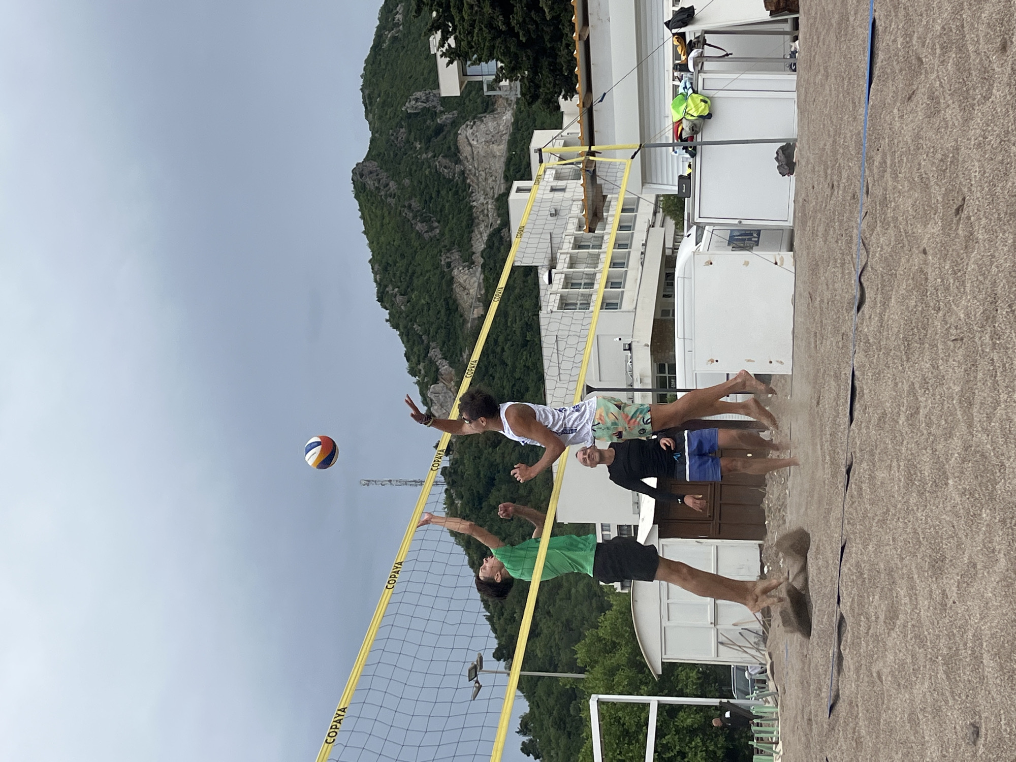 Three men are playing beach volleyball in Boreti, Montenegro. One man, wearing a green shirt and black shorts, is jumping at the net to block the volleyball. Another man, wearing a white tank top and patterned shorts, is jumping to hit the volleyball over the net. A third man, wearing a black long-sleeved shirt and blue shorts, is standing on the beach near the net, possibly waiting to receive the volleyball. The volleyball net is yellow and has the word "COPAYA" printed on it. The beach is sandy. In the background, there is a mountain, buildings, and cloudy sky.