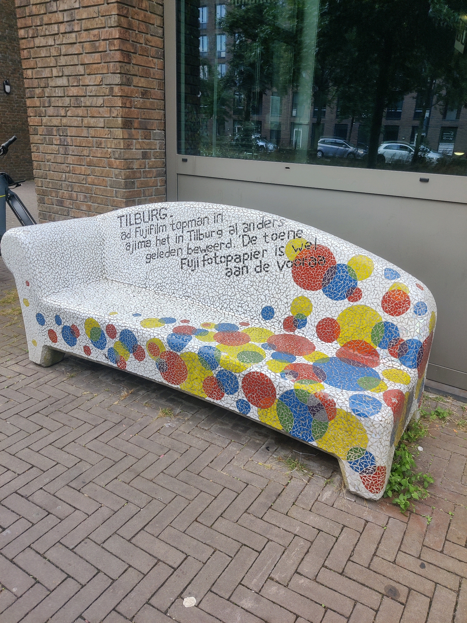 The image shows a mosaic-covered bench in front of a brick building with large windows. The bench is white with a mosaic pattern of small triangles covering its surface, and larger, colorful circles of red, yellow, blue, and green are scattered across the sides and front. The text on the bench reads: "TILBURG. ad FujiFilm topman in, ajima het in Tilburg al anders. geleden beweerd. De toene, Fuji fotopapier is wel aan de Voorla". This appears to be a reference to the Fujifilm factory and its history in Tilburg. The building facade includes brickwork and a large glass window reflecting trees and buildings. The ground is paved with gray bricks laid in a herringbone pattern. There is some grass and weeds growing around the base of the bench. A bicycle is partially visible on the left side of the image. The scene is set outdoors during the daytime.