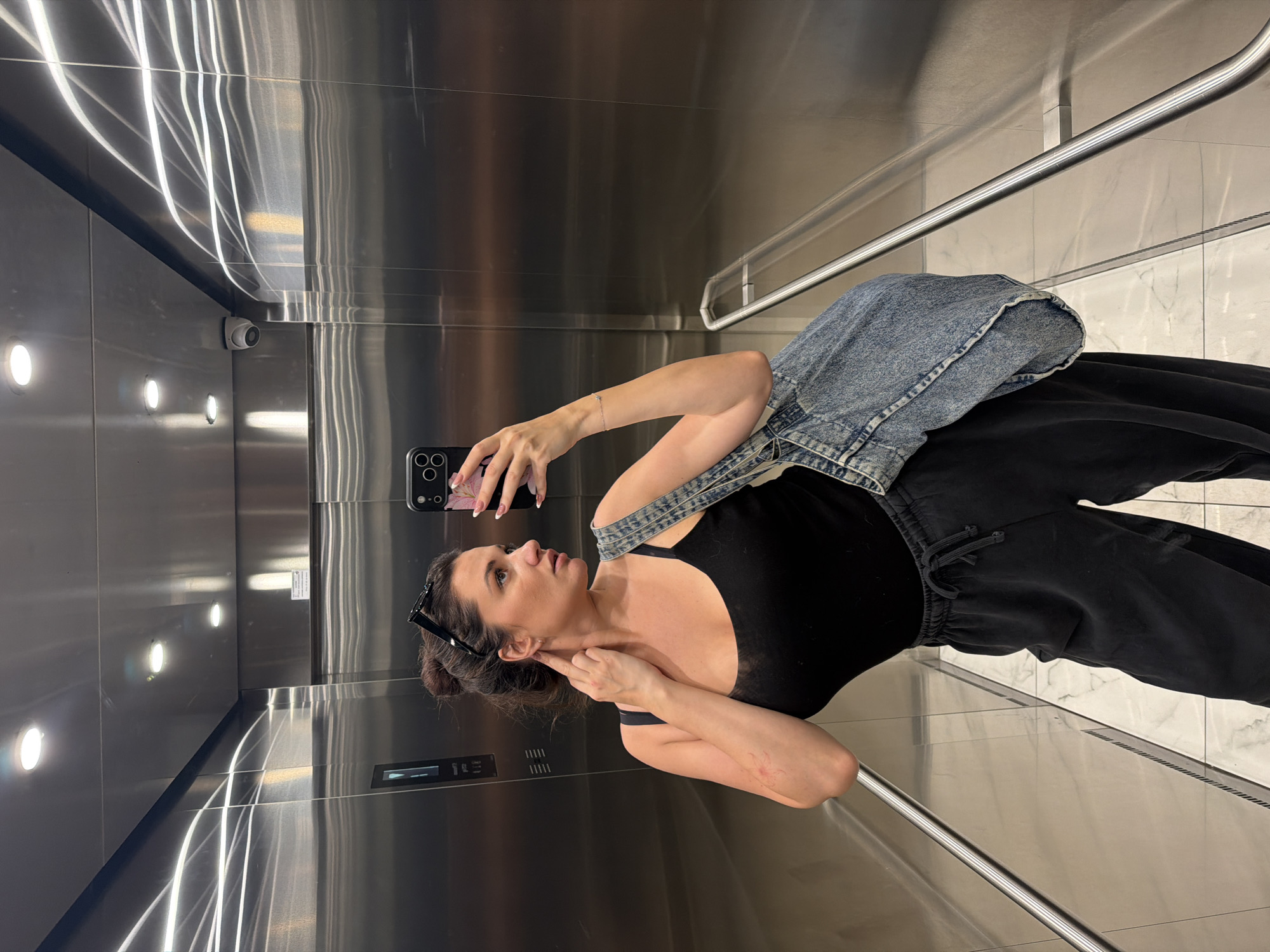 A woman takes a selfie in an elevator. The elevator interior is stainless steel, with a handrail. A security camera is visible in the upper left corner. The woman is wearing a black tank top, black sweatpants, and a denim jacket or bag. She is holding a smartphone with a floral case. The floor of the elevator appears to be marble or tile. The elevator has a display panel. The location is in an elevator in Belgrade, Serbia.
