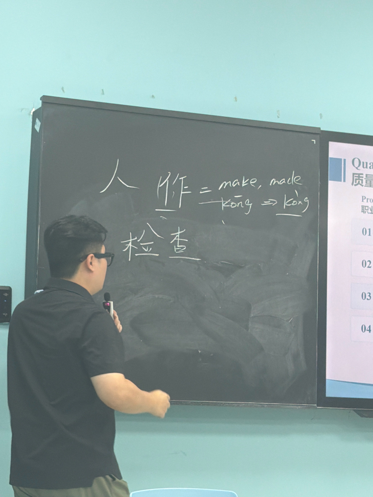 An Asian male, appearing to be in his 20s or 30s, wearing glasses and a black short-sleeved shirt, stands to the left of a blackboard in what appears to be a classroom. He holds a microphone in his right hand and is facing the blackboard, which has Chinese characters and English words written on it in white chalk. The characters "人" and "作" are written at the top, followed by the English "make, made." Below this, the word "kòng" is written, with an arrow pointing to "kòng" underlined. Further down on the blackboard are the characters "检查". To the right of the blackboard is a presentation screen with the title "Qua" and "质量" at the top, followed by "Prof" and then numbered items "01", "02", "03", and "04". The walls of the room are painted a light blue. The overall impression is that of a lecture or presentation in progress. The location is not discernible from the image itself.