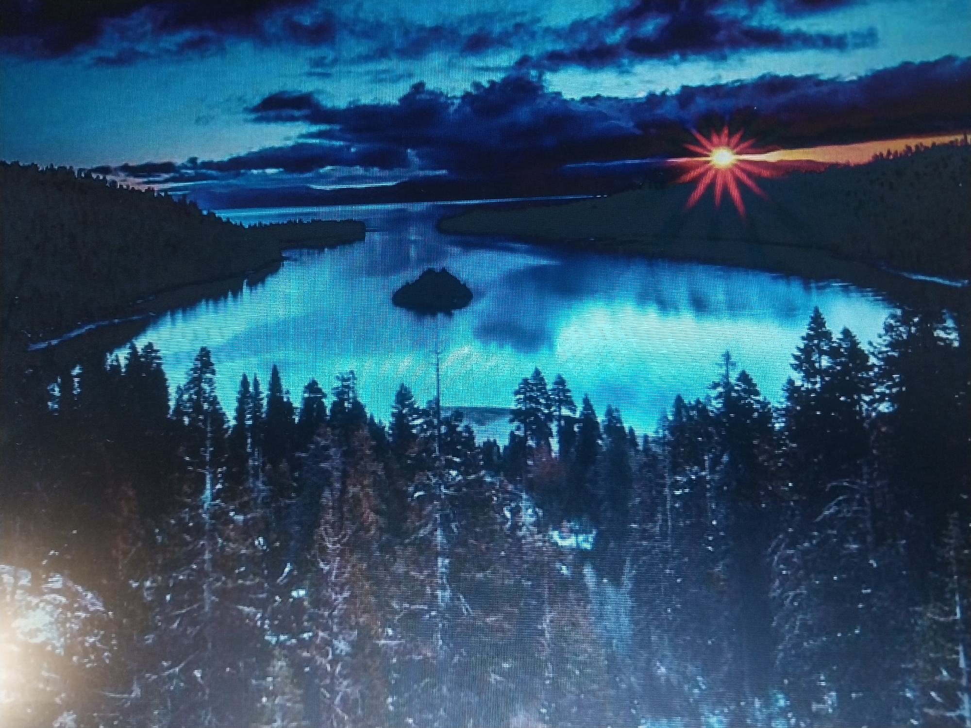 This is a high-angle, wide shot of a serene lake at dusk. The sky is a deep blue with dark, dramatic clouds, and the sun is setting on the right horizon, casting a warm, orange glow and radiating starburst rays. The water reflects the sky, appearing a brilliant blue. A small, dark island sits in the middle of the lake. The foreground is dominated by a dense forest of pine trees, some of which are dusted with snow, creating a wintery atmosphere. The overall impression is one of natural beauty and tranquility.