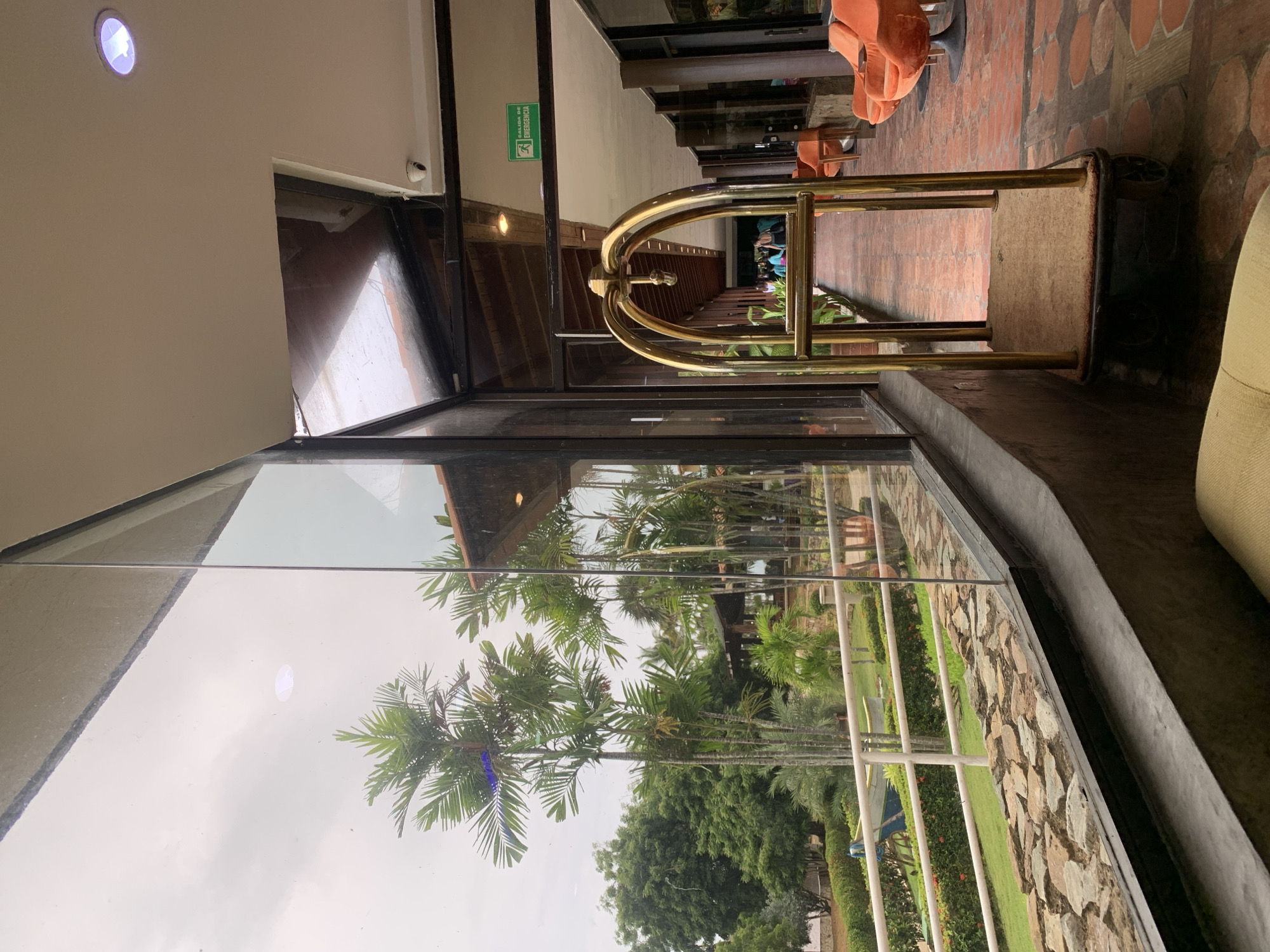 The image depicts an indoor hallway or lounge area with a large glass wall offering a view of an outdoor tropical landscape, situated in Playa El Agua, Venezuela. In the right midground, a golden-colored bellhop luggage cart with a beige base stands on a reddish-brown tiled floor. To the right of the cart, several orange, swivel-base armchairs are visible. The indoor ceiling, painted white, features recessed lighting and a small, white security camera mounted near a corner. A green "SALIDA DE EMERGENCIA" (Emergency Exit) sign is affixed to the ceiling structure further down the hall.
Through the expansive glass wall, an outdoor area is visible under an overcast sky. Prominent in the foreground of the outdoor scene are several tall, slender palm trees, likely Areca palms, with lush green fronds. Behind these palms, a variety of other tropical trees and manicured green bushes line a lawn. A white railing separates the immediate grassy area from more distant grounds where a blue and white boat, seemingly decorative, is positioned. Further in the background, a building with a reddish-brown tiled roof can be seen. Along the outdoor path, several indistinct figures, appearing to be people, are visible in the distance, suggesting activity in a resort-like setting. The base of the glass wall is constructed from rough-hewn stone tiles, transitioning to a smooth concrete ledge in the bottom left foreground, where part of a light-colored cushion or sofa is visible. Reflections of the indoor ceiling lights and structural elements are apparent on the glass surface.
