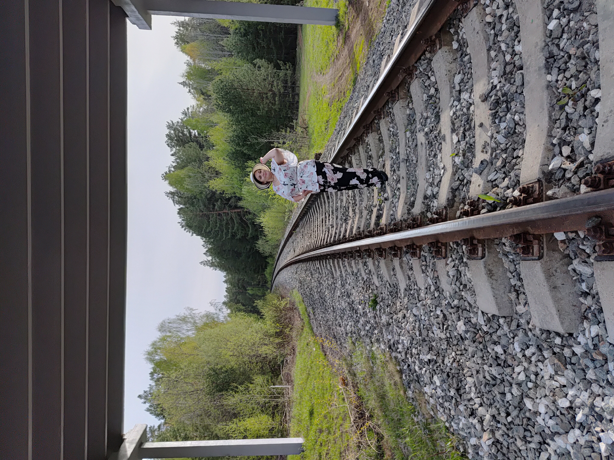 The image shows a woman in a hat, floral shirt, and black pants standing on railroad tracks. The tracks curve into the distance, surrounded by gravel and patches of green vegetation. A forest of green trees is visible in the background. To the left, there is a partial view of a modern structure, possibly a bridge or building, with a gray roof. The photograph appears to be taken in an outdoor setting. The image might have been taken in Chaikovsky, Russia, considering that city was mentioned in the prompt.