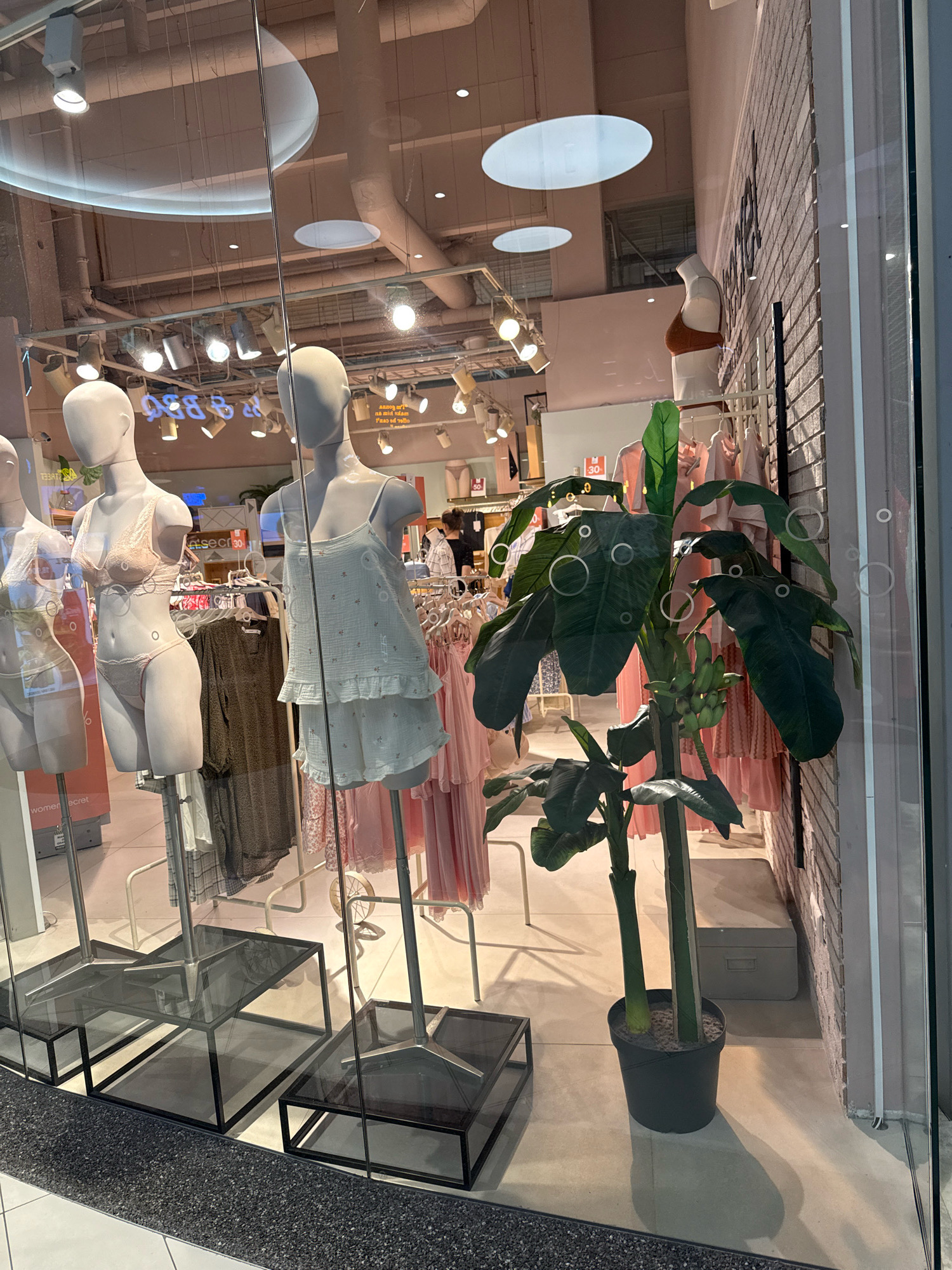 This image shows a retail store window display. Inside the window, several mannequins are dressed in lingerie and sleepwear. There is also a potted plant with large leaves and what appears to be bananas growing on it. Racks of clothing are visible behind the mannequins. The store appears to be a women's apparel store. The reflections in the window suggest an indoor shopping environment. The store is located in Tallinn, Estonia.