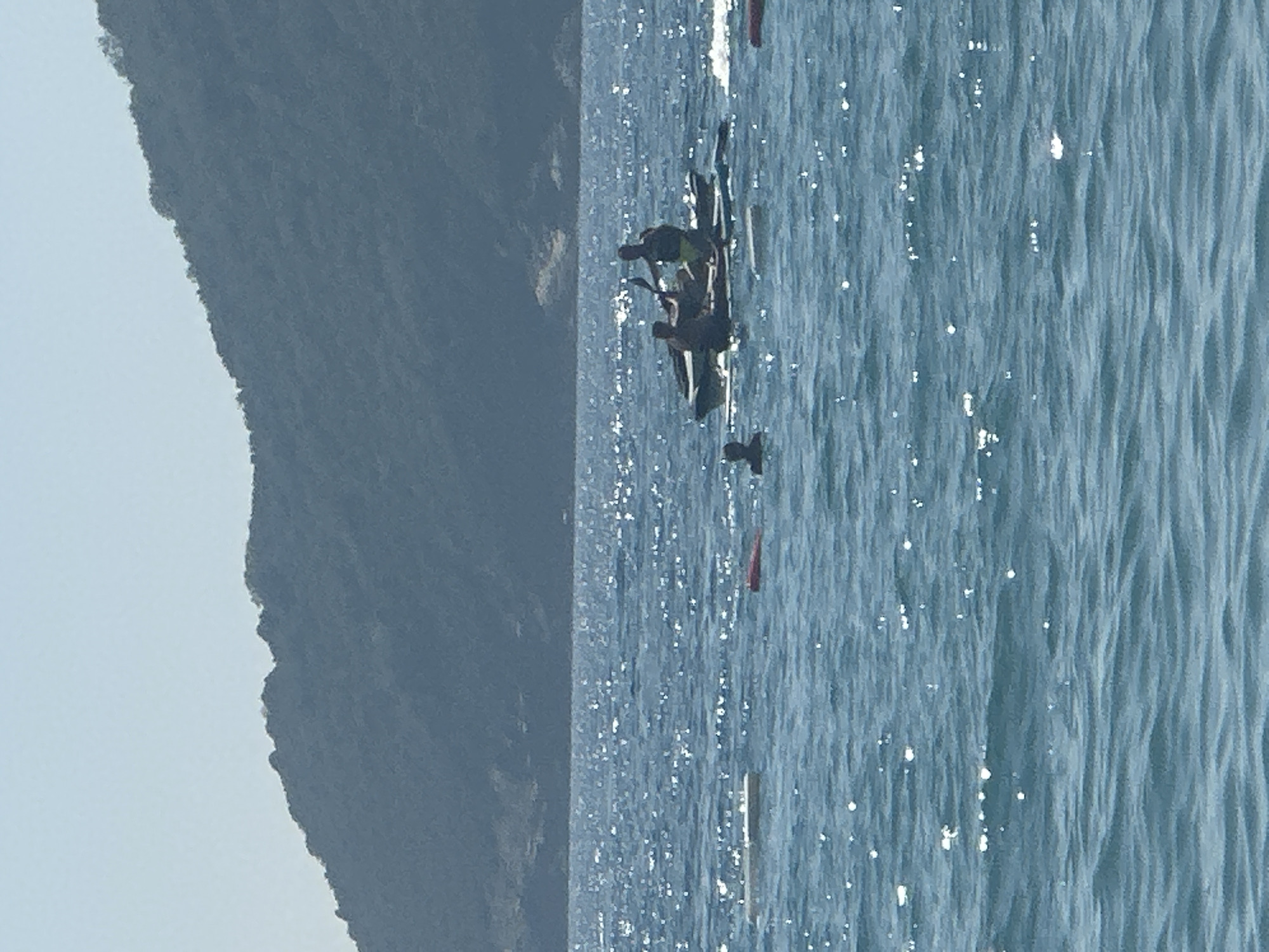The image depicts a body of water with a mountainous landscape in the background, likely the Bay of Kotor in Boreti, Montenegro. In the mid-ground, two individuals are seated in a kayak on top of a paddleboard. One person is holding a paddle. To the left of the kayak, a single person's head is visible above the water. Scattered throughout the water are several floating buoys or markers, one white and one red. The water is choppy and reflects sunlight. The sky is clear and pale blue.