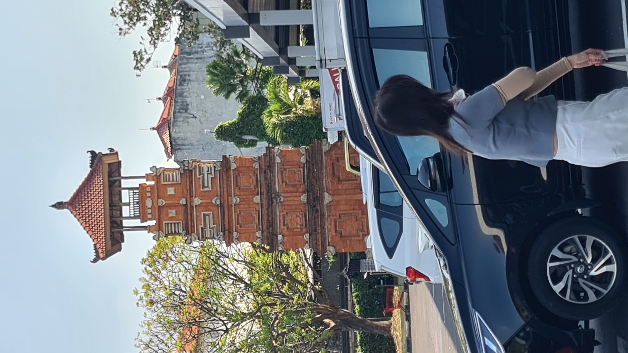 This image, captured in Denpasar, Indonesia, features a traditional Balinese tiered brick tower with a red tile roof, possibly a temple or a historical landmark. In the foreground, a woman with long dark hair is walking, wearing a grey short-sleeved top and light-colored pants. She is carrying a white bag. She is positioned next to a black car, partially obscuring a white car behind it. The scene is set outdoors, with trees and other buildings visible in the background, under a clear sky. The overall impression is of a sunny day in an urban environment with distinct cultural architecture.