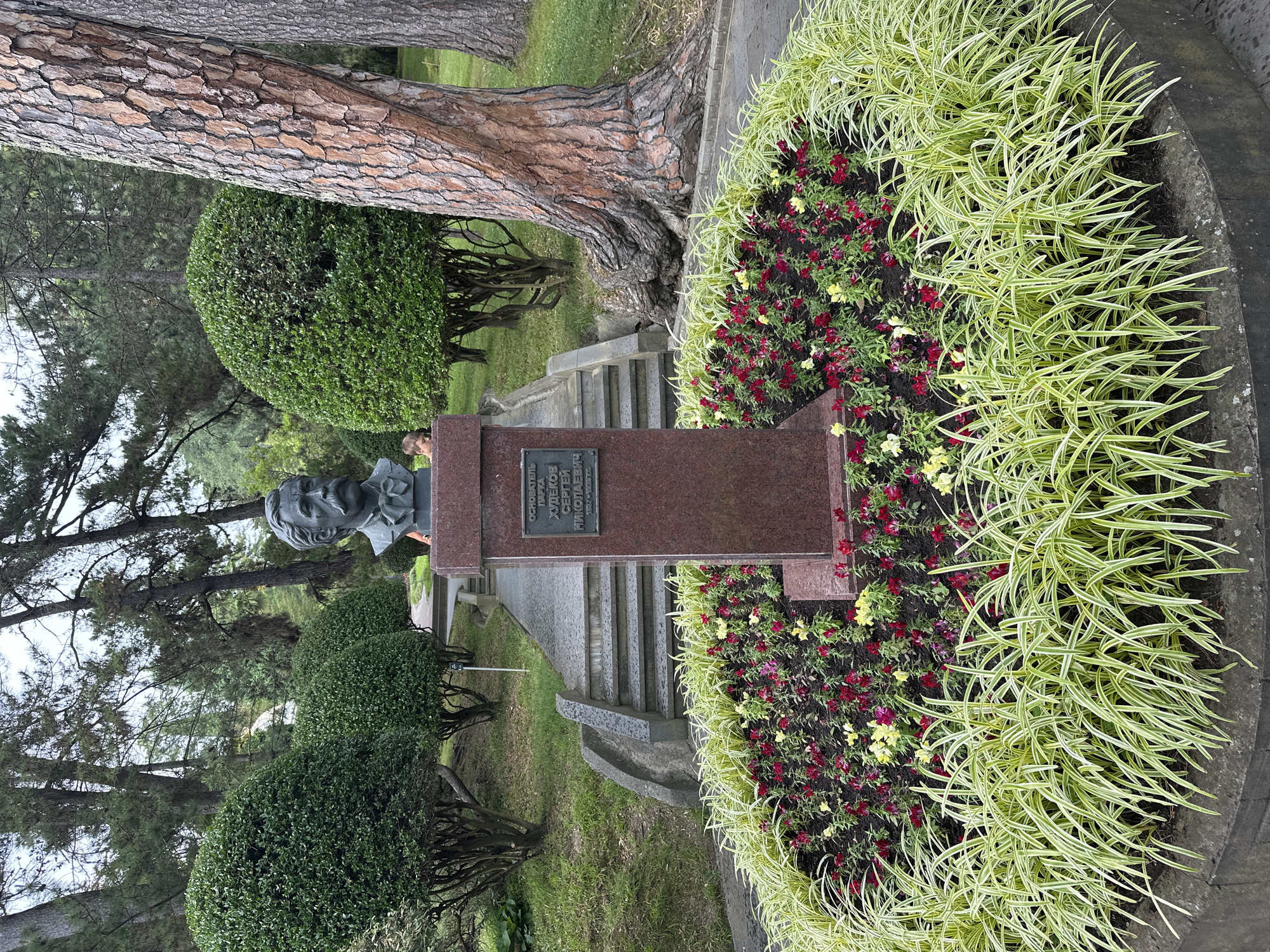 This is a medium shot photograph of a bust of a man with a mustache, located in a park in Sochi, Russia. The bust is made of bronze and rests on a red granite pedestal. A plaque on the pedestal reads: "ОСНОВАТЕЛЬ ПАРКА ХУДЕКОВ СЕРГЕЙ НИКОЛАЕВИЧ" (Founder of the Park Khudekov Sergey Nikolaevich). The bust is surrounded by a circular flower bed with red and yellow flowers and green grass. In the background, there are several trees and a paved path leading up a set of stairs. A young boy is visible standing behind the pedestal, looking down at the flowers. The park appears to be well-maintained with manicured bushes. The overall lighting suggests it is daytime, possibly overcast.