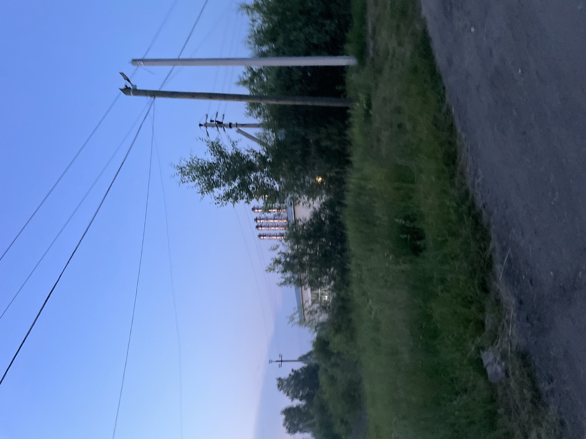 The image shows an industrial facility with multiple smokestacks in the distance. In the foreground, there is a dirt road on the right and a grassy area with tall grass and some bushes on the left. Several utility poles with power lines are visible across the scene, stretching into the blue twilight sky. The sky is clear with a gradient of light blue to dark blue, indicating dusk. The industrial building is light-colored with visible windows. The vegetation is dense and green. The overall scene depicts an industrial area on the outskirts of the city of Prudny, Russia, at twilight. There are no people or animals visible in the image.