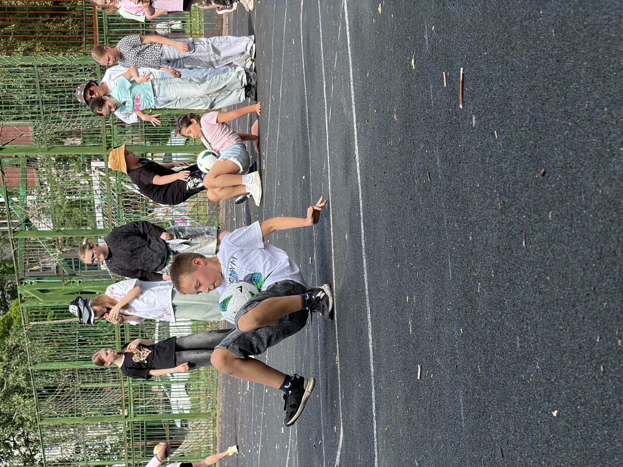 The image depicts an outdoor basketball court with a group of children engaged in some activity. In the foreground, a boy is performing a trick with a soccer ball, balancing it on his leg while in a seated position on the ground. He is wearing a white t-shirt and dark shorts. Behind him, a girl in a pink shirt is also on the ground, seemingly participating in the same activity with another soccer ball. Several other children and at least one adult are standing in the background, observing. The court is paved with a dark, textured surface with white lines. A green metal fence is visible behind the group, and some trees and foliage can be seen beyond the fence. The setting appears to be a public park or recreational area. There is no specific indication of this location being in Izhevsk, Russia; it is a general outdoor court.