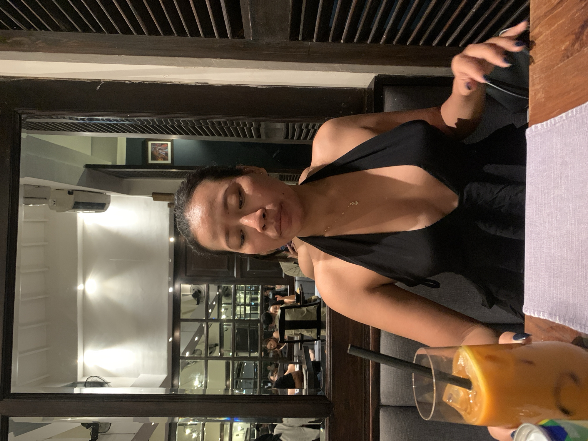 A woman sits at a table in a Kuta, Indonesia, establishment. She wears a black halter top and a delicate gold necklace. In front of her is a glass of iced coffee with a straw, and her hand rests near the edge of a white cloth. The background features a series of mirrors reflecting the restaurant's interior, showing other patrons seated at tables. The architectural details suggest a restaurant or cafe setting.