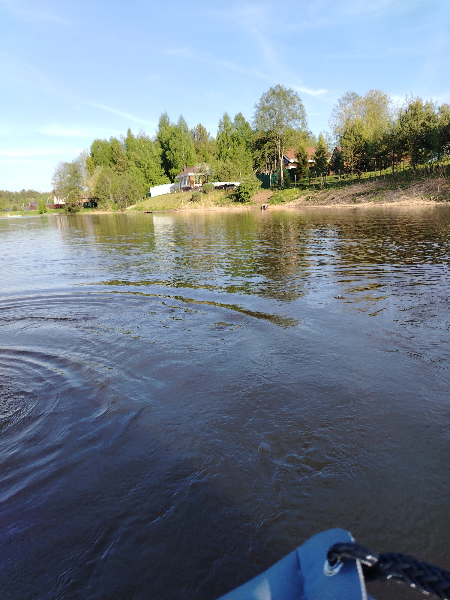 The image shows a body of water, likely a river, with visible ripples and dark coloration. The water extends into the foreground, where a portion of a blue object with a black rope is visible. Along the far bank of the water, there is a strip of land with green trees and vegetation. Several buildings, possibly houses or cabins, are visible nestled amongst the trees. The sky above is a light blue with a few scattered clouds. Based on the location of Stroitel', Russia, the scene could be a section of a river in a rural or semi-rural area near the town.