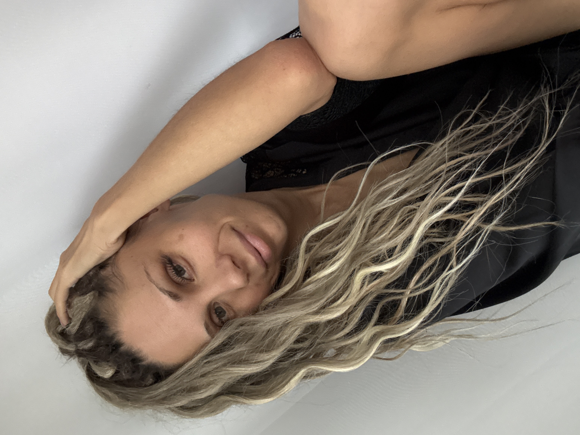 A woman is sitting with her arm raised to her head. She is wearing a black robe with lace detailing. Her hair is long and blonde, with some darker roots. She is looking towards the camera with a slight smile. The background is a plain white wall, and there is a hint of white fabric in the foreground. The image appears to have been taken indoors, with natural light. There is no discernible indication of the location in the city of Rusanovo, Russia.