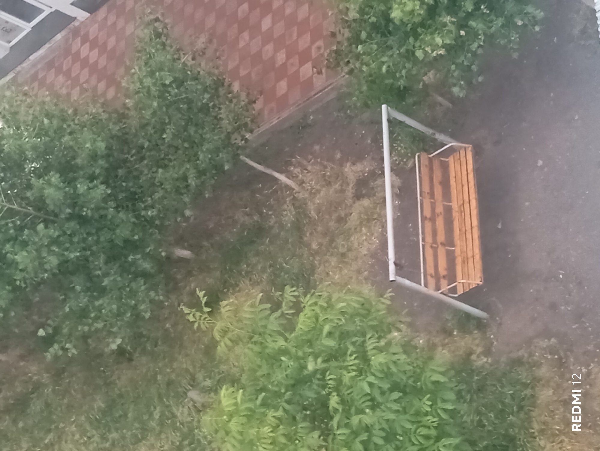 The image displays an outdoor scene, likely a residential courtyard or communal garden, viewed from an elevated perspective. The central subject is an empty, two-person swing set featuring a light-colored metal A-frame and a wooden slatted seat with a light brown-orange finish.
The ground immediately surrounding the swing set is dark, appearing as bare earth or sparse, dry vegetation. To the left and upper left, a dense area of vibrant green foliage, including bushes and small trees with varied leaf shapes, occupies a significant portion of the frame. Towards the upper right, more green foliage is visible, interspersed with patches of lighter, dry ground, where a fallen branch or stick also lies.
In the upper right quadrant, a section of a paved area is present, characterized by a clear checkerboard pattern of reddish-brown and lighter square tiles. Adjacent to this paved area, along the top right edge, is the lower section of a building with a dark grey or black base wall, topped by partially visible white-framed windows.
The scene is uniformly illuminated, suggesting an overcast sky or diffuse light conditions, as no harsh shadows are apparent. No individuals are visible in the frame. A "REDMI 12" watermark is located in the bottom left corner of the image. The location is in the city of Unknown, Unknown.