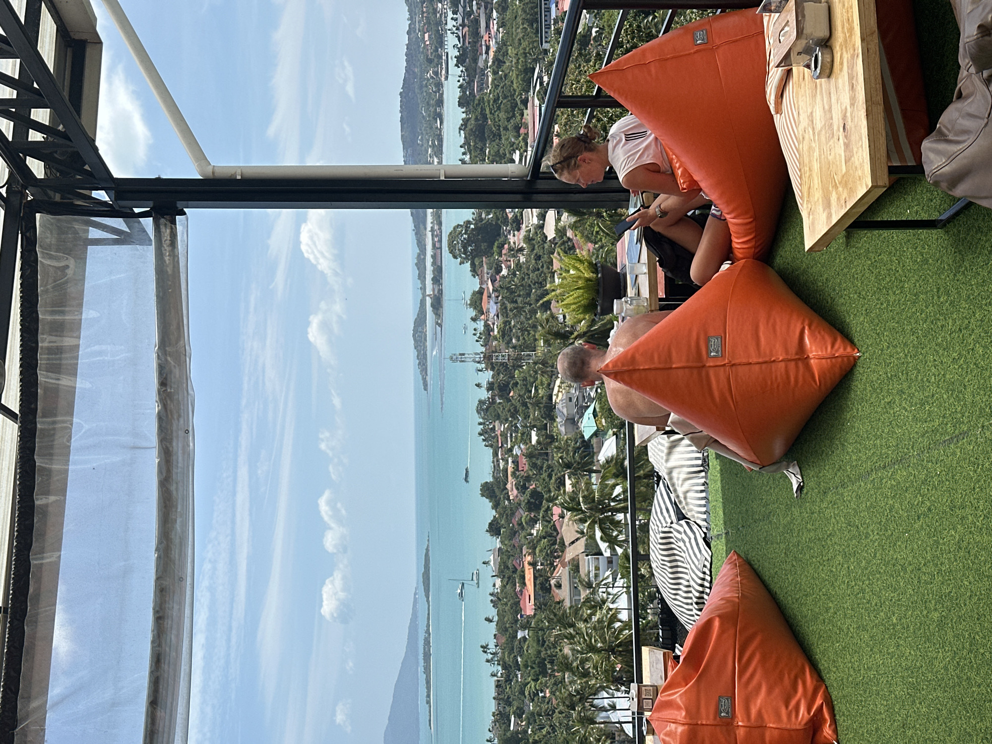 In the city of Ko Samui District, Thailand, this image captures a scenic view from an elevated outdoor seating area. Two individuals are seated on orange beanbag chairs, facing the camera with the backdrop of a vibrant coastal landscape. To the left, a man with a shaved head is partially visible, leaning back. To the right, a woman with blonde hair pulled back is looking down at her mobile phone. In front of them, a wooden table holds a potted plant and a glass of water. The seating area is covered by a transparent roof supported by a metal frame, offering protection from the elements while allowing ample natural light. Beyond the seating area, a sprawling view of Ko Samui unfolds, with lush green hills dotted with houses and palm trees leading down to a turquoise ocean. Several boats are visible on the water, and islands can be seen in the distance under a bright blue sky with scattered white clouds. The artificial green turf flooring of the seating area contrasts with the natural beauty of the surrounding environment.