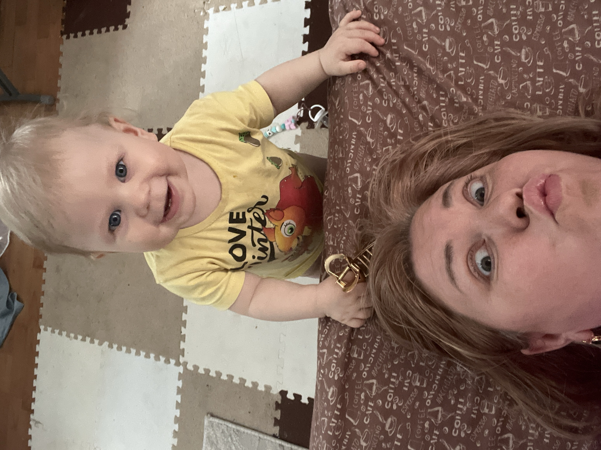 A selfie captures a woman and a child. The woman, on the right, has light brown hair, fair skin, and is making a kissing face toward the camera. To her left, a young child with fair hair and blue eyes smiles, wearing a yellow t-shirt with a graphic print and the words "LOVE inter."
The background is a brown patterned surface with coffee-related words printed on it, possibly a bed or a cushion. To the left, the floor is covered with interlocking foam play mats in a checkered pattern of light and dark squares. There are no discernible landmarks or features indicating a specific location in Vesyoloye, Russia. The focus is primarily on the people in the image.