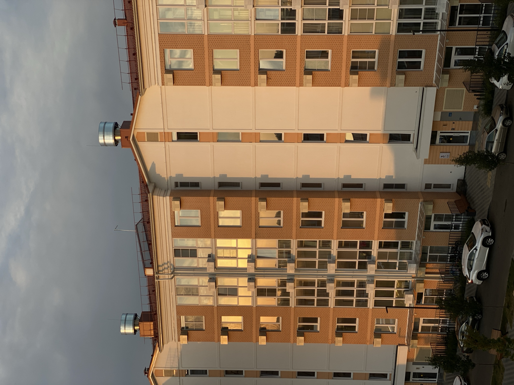 The image depicts a multi-story residential building with a brick facade, viewed from a slightly elevated perspective. The building features numerous windows with reflective glass, some of which show the warm glow of sunset. Various air conditioning units are mounted on the exterior walls. On the rooftop, there are several ventilation systems or chimneys, one of which is a large cylindrical metal unit. The sky above is partly cloudy with darker, heavier clouds dominating the upper portion. In the foreground, a street or courtyard is visible with parked cars, including a white SUV and a dark sedan, and some pedestrian pathways. Trees and benches are also present. The scene appears to be an urban residential area, likely Dinskaya, Russia, as suggested by the architectural style and context of similar buildings. No people are clearly visible. The lighting suggests it is late afternoon or early evening.