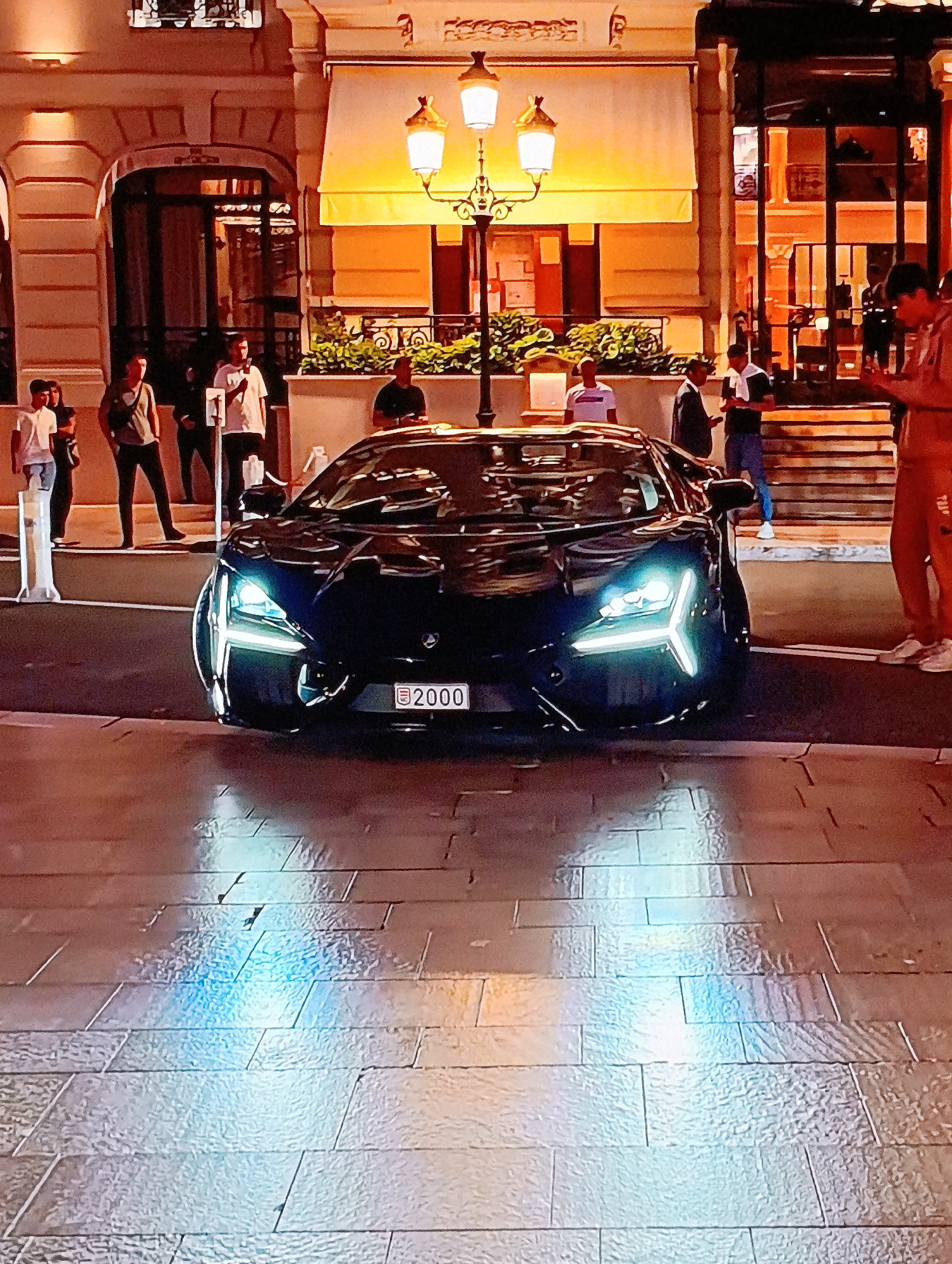 The image shows a dark-colored, high-end sports car parked on a cobblestone street at night. The car has distinctive, illuminated headlights that form a "Y" shape. It features a license plate. Behind the car are several people standing and walking. The background includes a building with illuminated awnings, ornate architectural details, and a lamppost with multiple lamps. The light from the car's headlights reflects off the cobblestone, creating bright, blue-white streaks on the ground. The location appears to be a city street in Monaco.