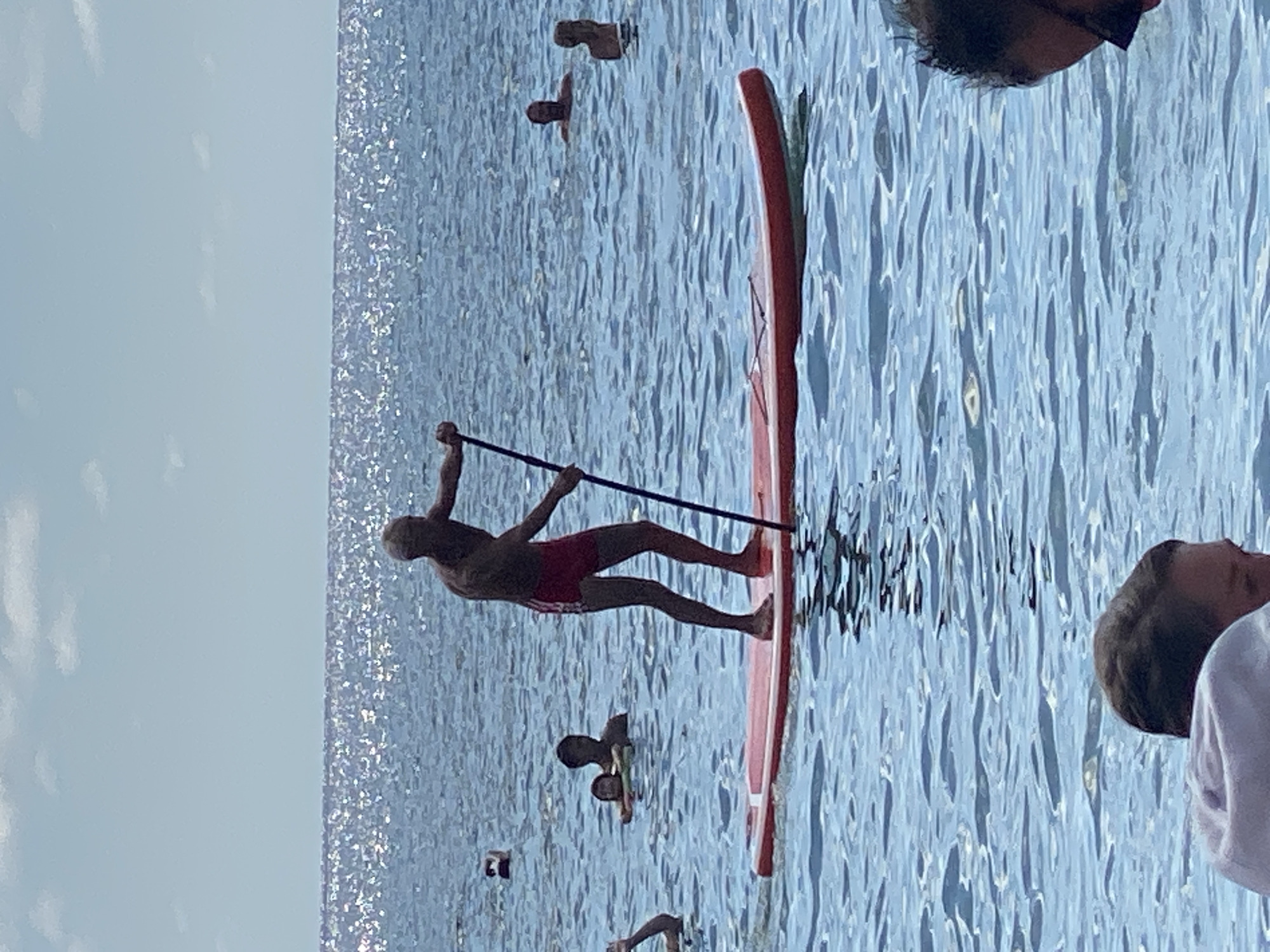 A full shot, eye-level, medium-distance view captures a person stand-up paddleboarding on a body of water. The paddleboarder, silhouetted against the bright, sunlit sea, is wearing red shorts and is holding a paddle. The sea is a deep blue with sparkling sunlight reflecting off its surface. Other individuals are visible in the water further in the background. The scene appears to be in Arkhipo-Osipovka, Russia, a coastal resort town.