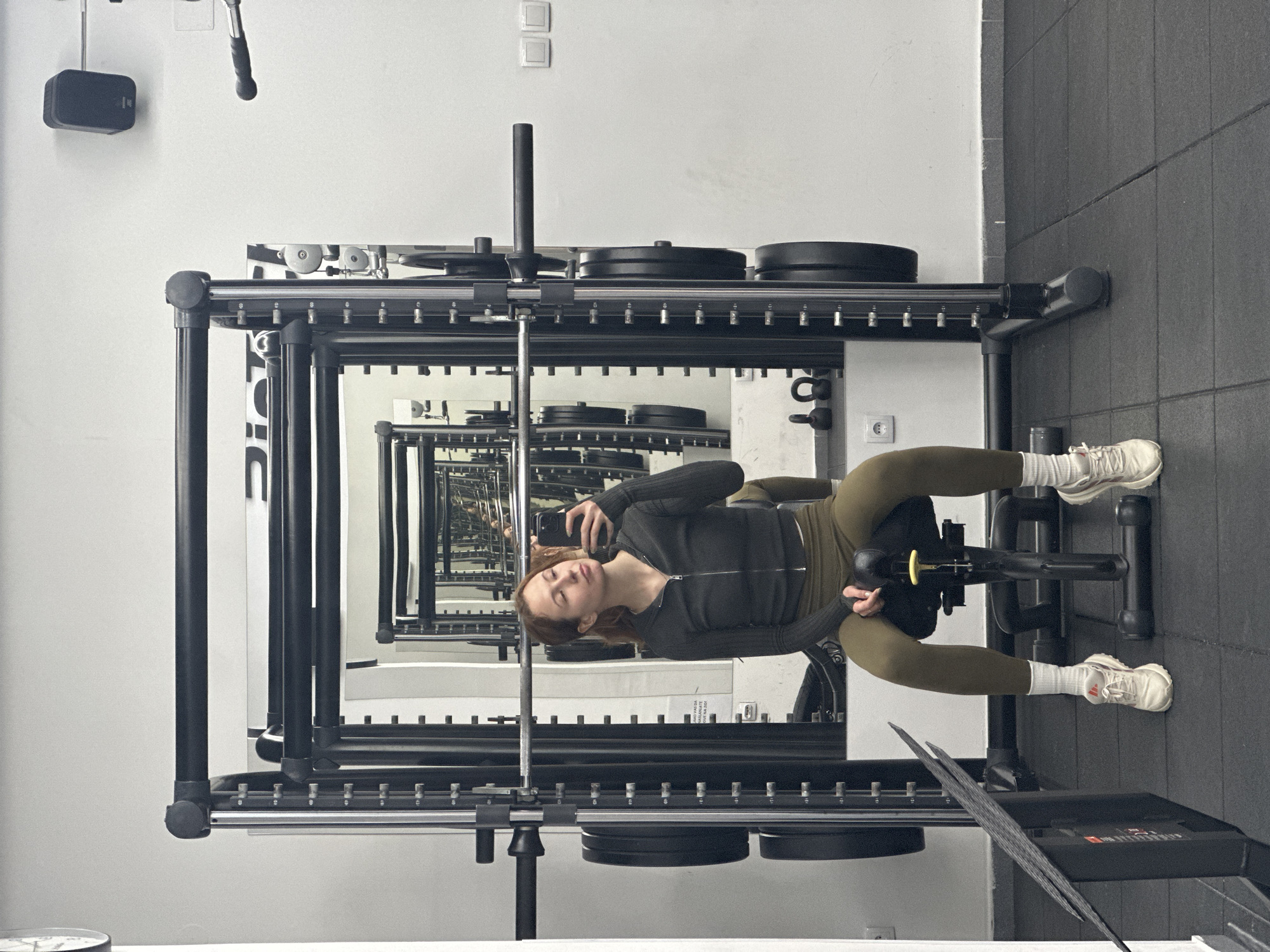 A person sits on a leg extension machine in a gym with mirrored walls, creating a repeating perspective. The person is fair-skinned with reddish hair, wearing a dark zip-up jacket, and olive-green leggings, with white sneakers and socks. They are holding a mobile phone, presumably taking a selfie. The gym equipment includes a weight rack with weights on it, and the floor is covered with black rubber tiles. The location is Novi Sad, Serbia, as indicated by the "NS Fit" logo visible in a cropped image.