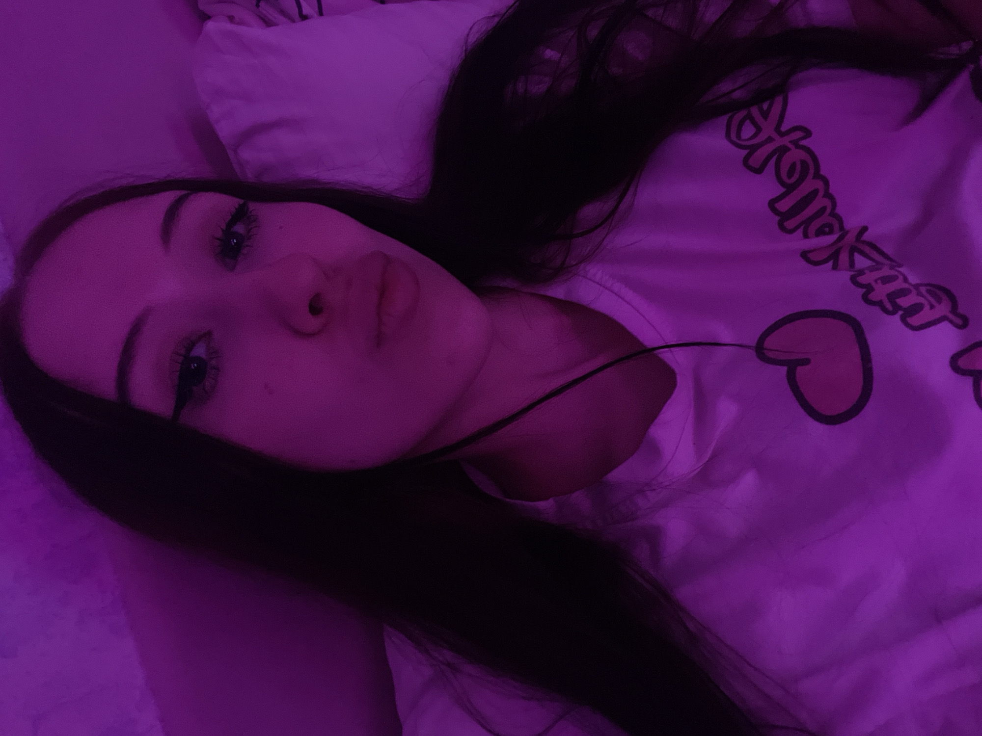 The image shows a close-up, overhead shot of a young woman lying down. She has long, dark hair and is wearing a white t-shirt with a graphic design. The lighting is purple, creating a moody atmosphere. The woman's face is partially visible, with her lips puckered as if for a kiss. Her eyes are open and looking towards the camera. The background appears to be a bed with white pillows. There is no indication of the location in Votkinsk, Russia.
