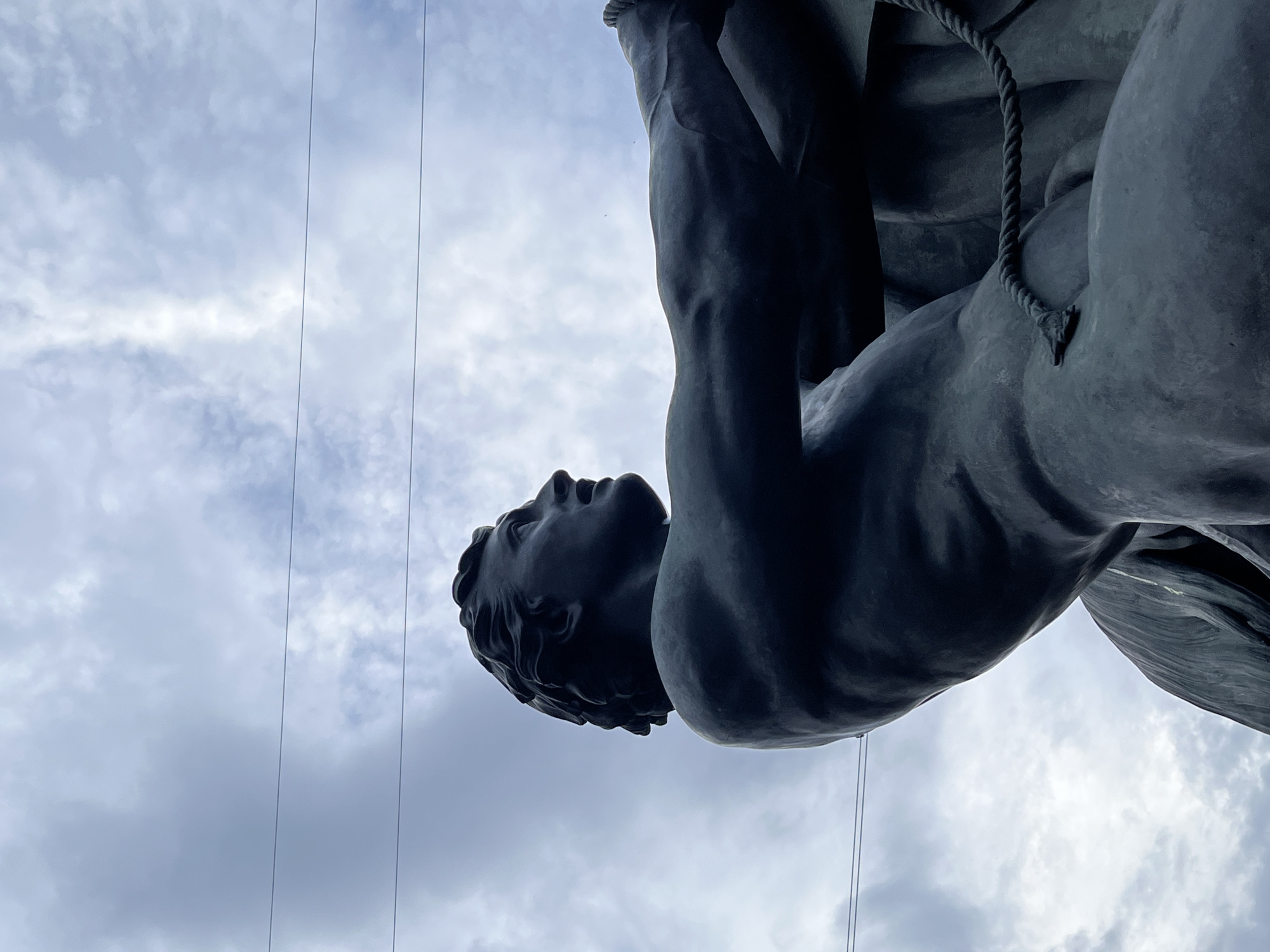 The image presents a low-angle, upward-looking perspective of a dark bronze statue of a muscular male figure silhouetted against a cloudy sky. The statue, depicting a man from the waist up, is positioned in profile, looking towards the upper right of the frame with a determined expression. His head is turned, showing short, wavy hair. His bare arms are extended forward and to the right, engaged in an action that suggests pulling or restraining. A dark rope is visible, wrapped around his lower torso and extending out of view to the right, reinforcing the sense of active engagement. The surface of the statue shows sculptural details of musculature.
The background consists of a partially overcast sky with varying shades of light blue and grey clouds. Two thin, dark utility wires horizontally traverse the upper portion of the sky, passing behind the statue. The overall lighting casts the statue in a deep shadow, emphasizing its form against the brighter sky.
This statue is one of the four "Horse Tamers" (also known as "Horse-Rapplers" or "Klodt's Horses") sculptures located on the Anichkov Bridge over the Fontanka River in Saint Petersburg, Russia. The specific pose with the visible rope is characteristic of these monumental works by Baron Peter Klodt von Jurgensburg.