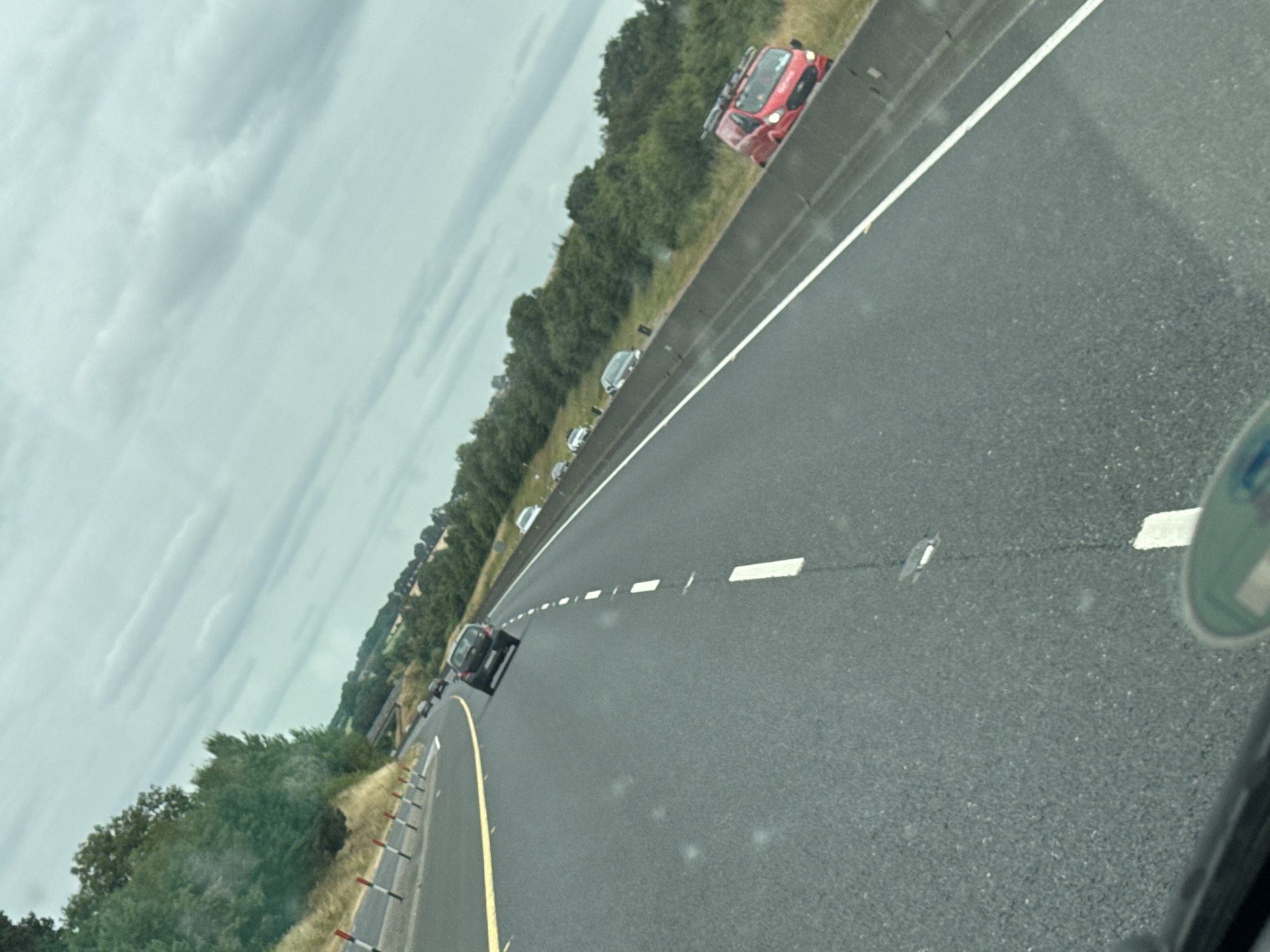 The image is taken from inside a vehicle traveling on a highway, looking towards the right and slightly upwards. The highway has multiple lanes and is paved with asphalt. A yellow line marks the edge of the lane, and white dashed lines separate the lanes. On the right side of the road, a concrete barrier separates the highway from a grassy verge with trees and bushes. Several vehicles are visible on the highway, including a dark-colored car in the foreground, a red van with a ladder rack on its roof parked on the verge, and other cars further in the distance. The sky is overcast with grey clouds. The landscape suggests a rural setting. The scene is likely a highway in or near Coolavash, Ireland, given the context provided.