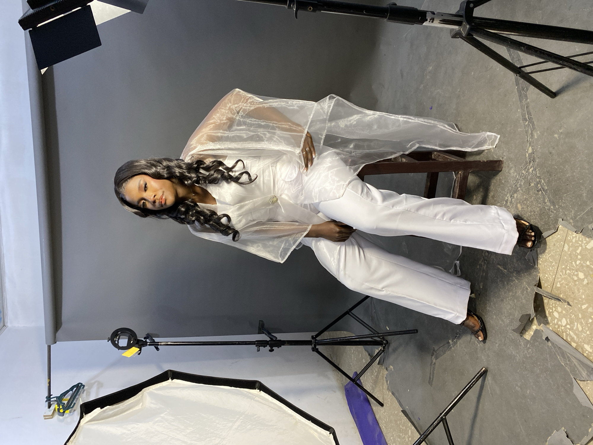 A woman poses for a photo in a studio setting in Jos, Nigeria. She is seated on a wooden chair, wearing a white suit with a sheer white overcoat and black sandals. The backdrop is gray, and various pieces of photography equipment, including lighting stands and softboxes, are visible around her.