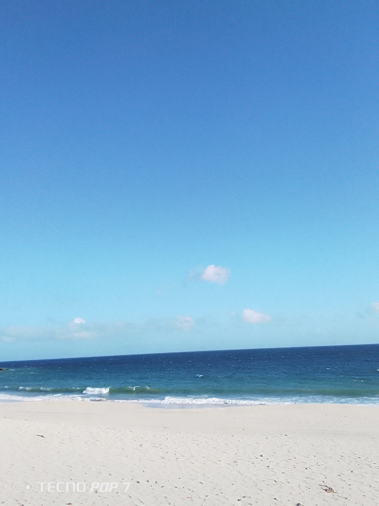 The image captures a serene beach landscape under a clear, bright blue sky, likely during daytime. In the foreground, a wide expanse of light-colored, possibly white, sand stretches across the bottom portion of the frame. The sand shows natural textures and a few scattered dark specks.
The midground features the ocean, displaying a deep blue hue that transitions to a lighter, turquoise-green closer to the shore. Gentle waves with white foam crests are seen breaking softly onto the sandy beach. The horizon line is straight and clearly defined.
Above the ocean, the background consists of a vast, unblemished blue sky, with only a few sparse, small, white, puffy clouds scattered towards the top. The overall atmosphere is one of peace and natural beauty, suggesting calm weather conditions.
In the bottom left corner of the image, a visible watermark reads "OO TECNO POP 7". The scene depicts a tranquil coastal view, consistent with a beach environment in Pedro González, Venezuela. No people or specific man-made structures are visible within the frame.