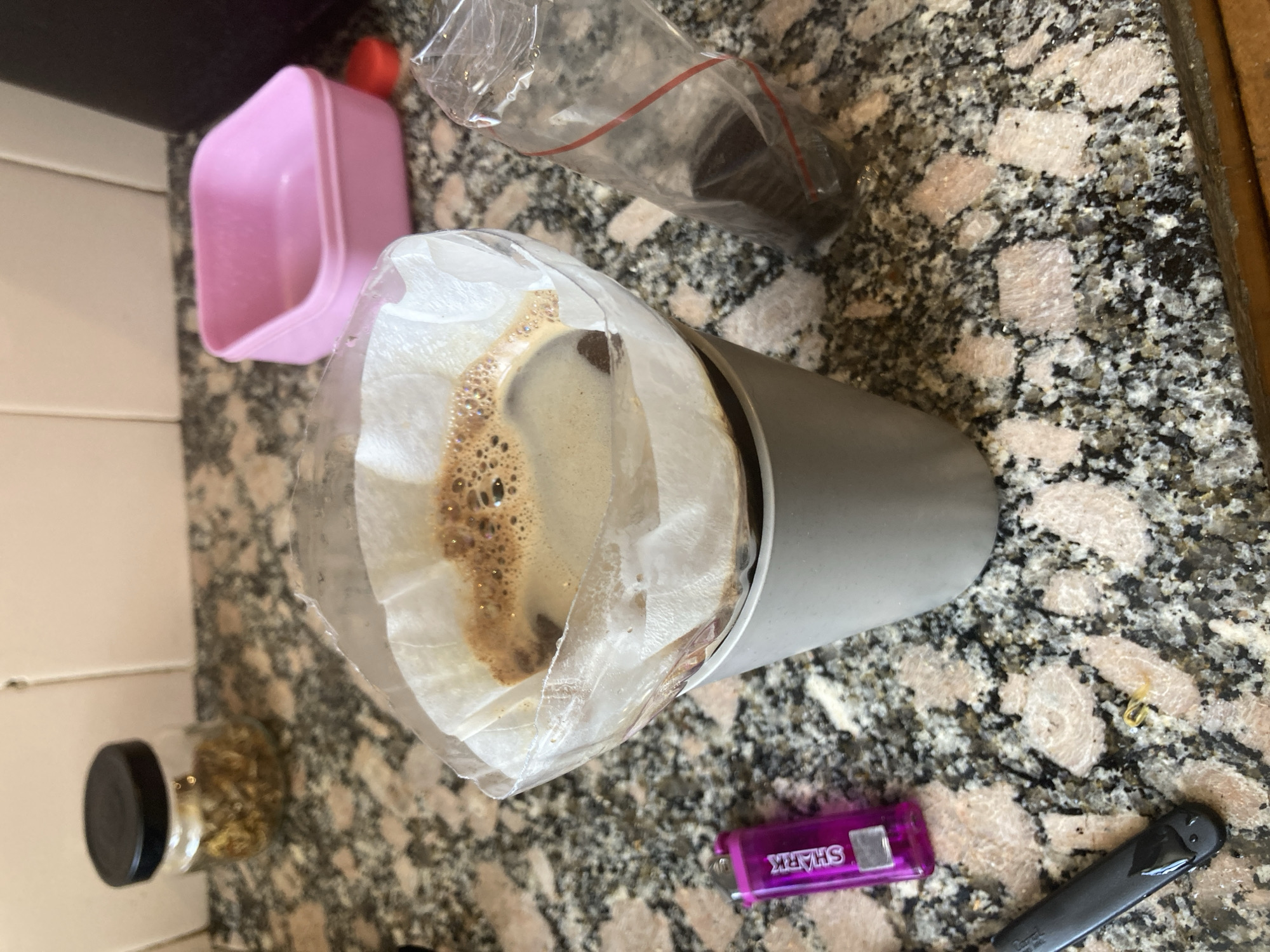 On a speckled countertop, likely in a kitchen, sits a grey cylindrical container with a coffee filter inserted, containing coffee grounds and brewed coffee. To the left is a small glass jar with a black lid filled with an unknown substance. Nearby, a pink plastic container is also visible. A clear plastic bag lies partially open, revealing a black object within. A purple lighter and a black tool are situated near the base of the coffee container. The environment suggests a preparation or disposal process related to coffee brewing. The specific location and city are unknown based solely on this image.