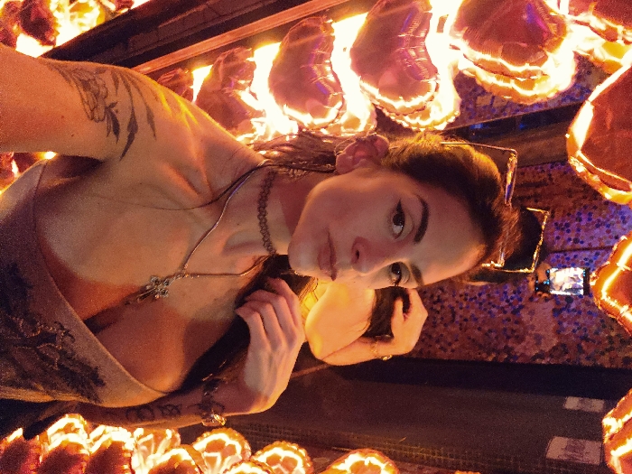 A female subject with dark hair, dark eyeliner, dark eyebrows, a labret piercing, and multiple ear piercings faces the camera directly with a neutral expression. Sunglasses are perched on her head. She wears a black choker, a silver necklace with a cross pendant, and a light-colored, low-cut, off-shoulder top. Tattoos are visible on her left upper arm, including a tribal-style design and a floral or leaf-like image below it. Her right hand is positioned behind her head, near her hair. The background is an interior space with a mosaic-like wall pattern in purple and blue tones. Numerous heart-shaped, illuminated objects, emitting an orange-red glow, surround the woman. These objects are arranged vertically along the left side and partially on the right. In the upper right background, a smartphone is visible, held as if capturing an image. The environment suggests an entertainment venue. The specific location within Buenos Aires, Argentina, cannot be determined from the visual information provided.