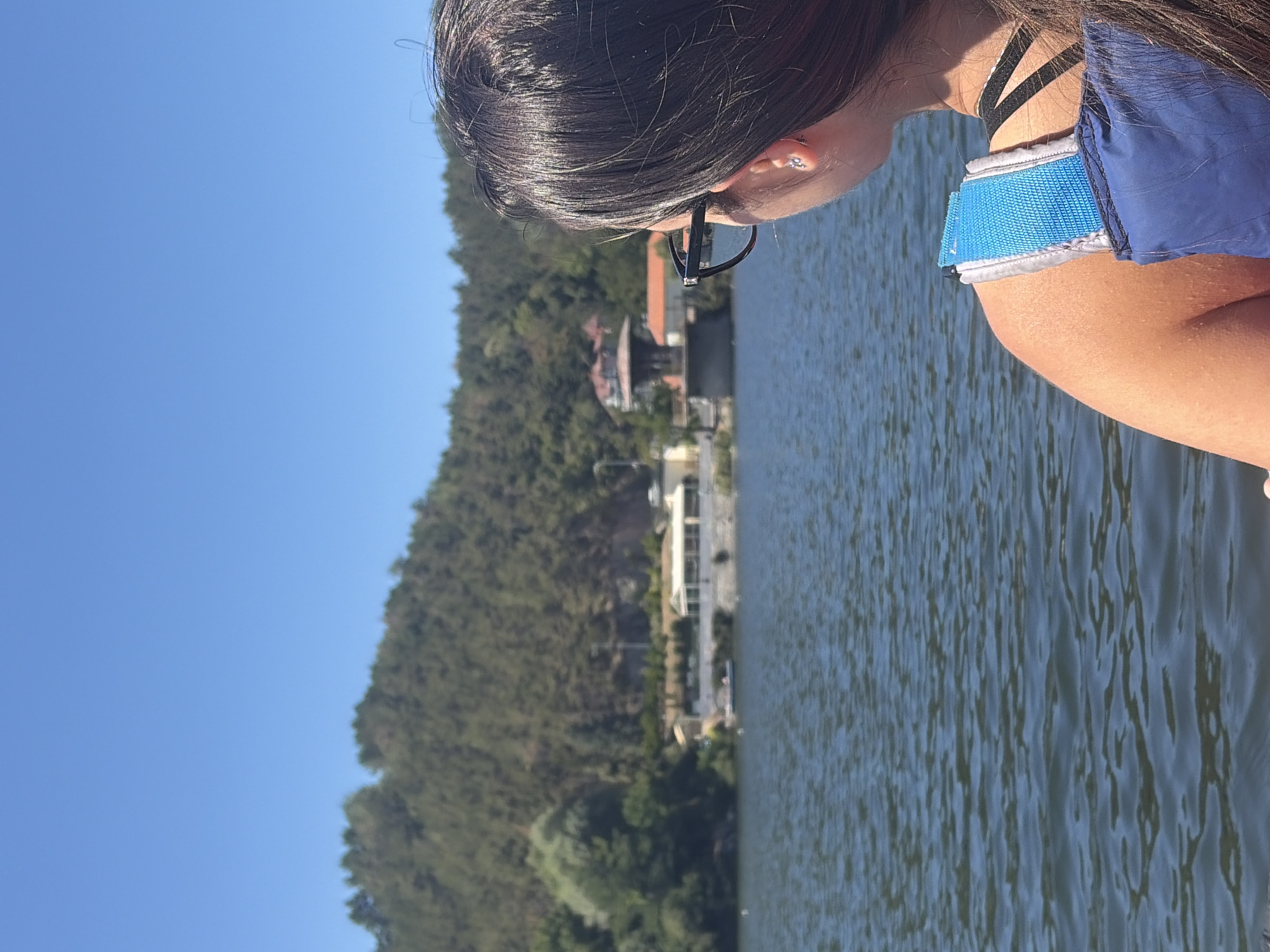 The image captures a view from a body of water, looking towards a tree-covered hillside with buildings nestled amongst the trees. In the foreground, a person's right shoulder and the back of their head are visible, as they wear a blue life vest over dark clothing. The person has long, dark hair and is wearing dark-rimmed glasses. The water is rippling, reflecting the clear blue sky above. The scene appears to be a sunny day. The location is identified as Pancharevo, Bulgaria.