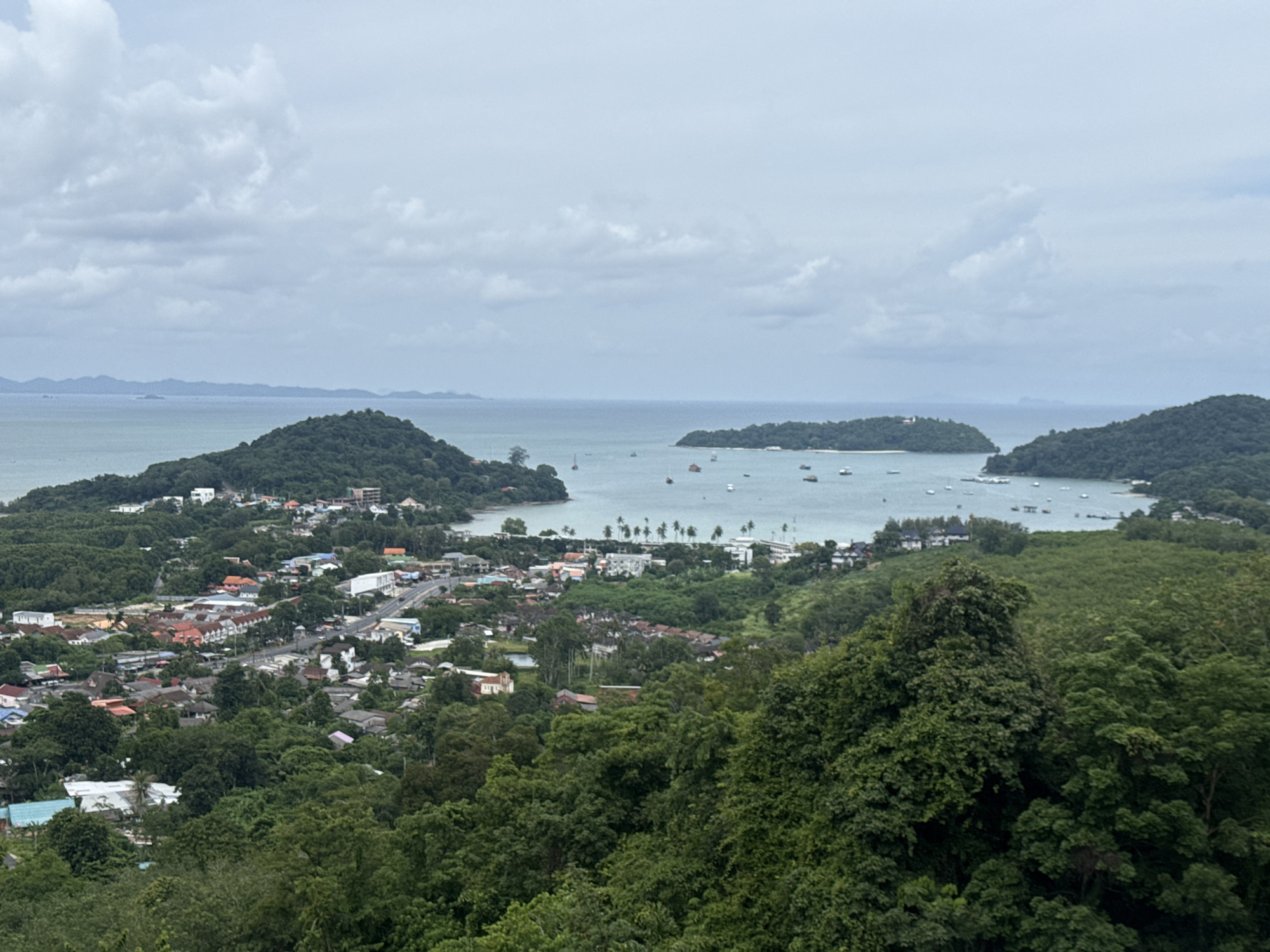 This is a high-angle, wide shot of a coastal town and bay. In the foreground, dense green foliage of trees obscures the lower portion of the image. Beyond the trees, a town with a variety of buildings, including residential homes with red and orange tile roofs, stretches towards the water. A paved road runs through the town, with several vehicles visible. The bay is dotted with numerous boats, varying in size, some appearing to be sailboats or yachts. Several tree-covered islands or peninsulas are situated in the bay, with one in the middle distance featuring a white building or structure. The water is a calm, muted blue-green, and the sky is overcast with scattered white and gray clouds. The overall scene suggests a moderately populated coastal community in a tropical or subtropical region. This appears to be a view of Chalong Bay, a popular tourist and boating area in Wichit, Thailand.