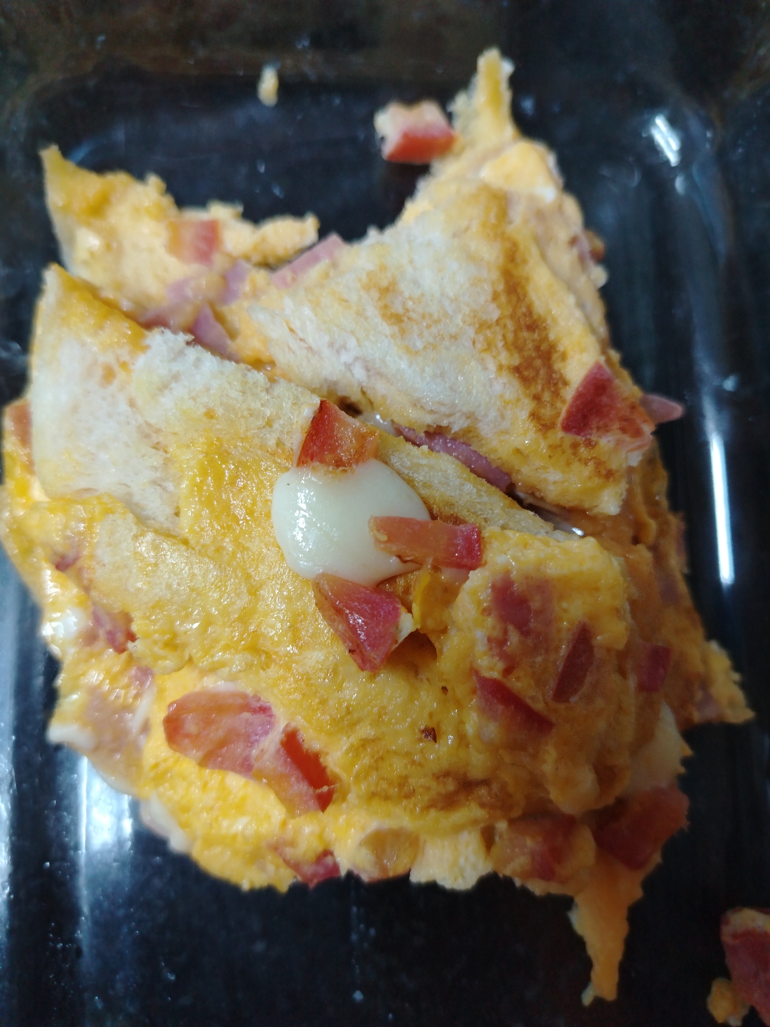 A close-up, overhead shot shows a portion of a sandwich cut in half, resting in a black plastic container. The sandwich appears to be made with white bread and is filled with scrambled eggs, diced red pieces that could be tomato or ham, and some melted white cheese. The bread has a grilled appearance with golden-brown stripes. The container is dark and slightly reflective, suggesting a plastic material. There are no visible people or specific landmarks indicating a location in Dubai, United Arab Emirates. The lighting is somewhat dim, creating a slightly grainy texture in the image.